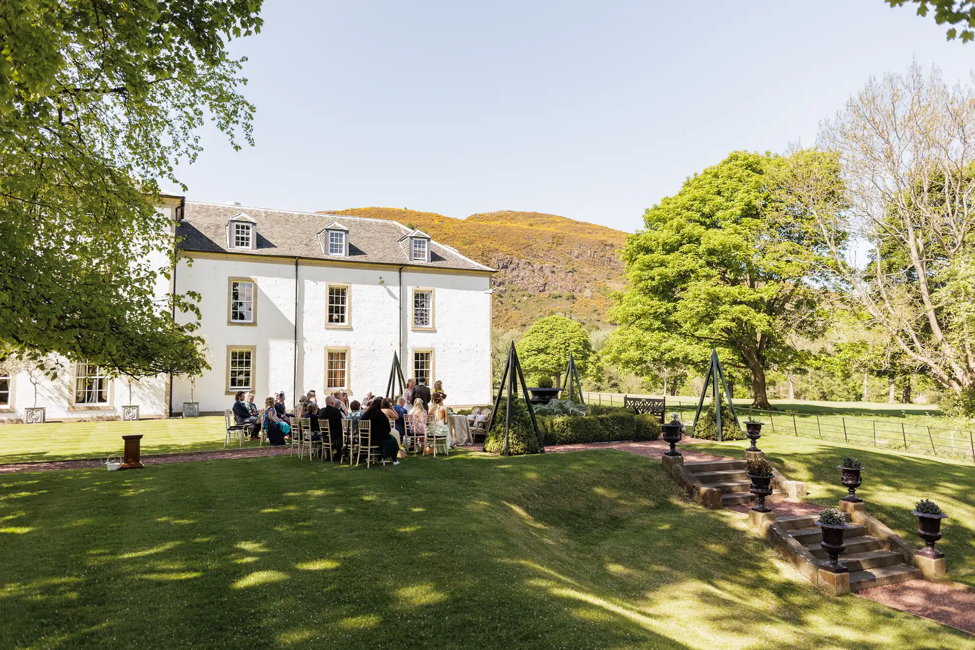 A group of people sits on chairs arranged in rows outside a large white house, attending an outdoor event. The scene is set on a sunny day with lush green grass, leafy trees, and hills in the background. The atmosphere is calm and picturesque.