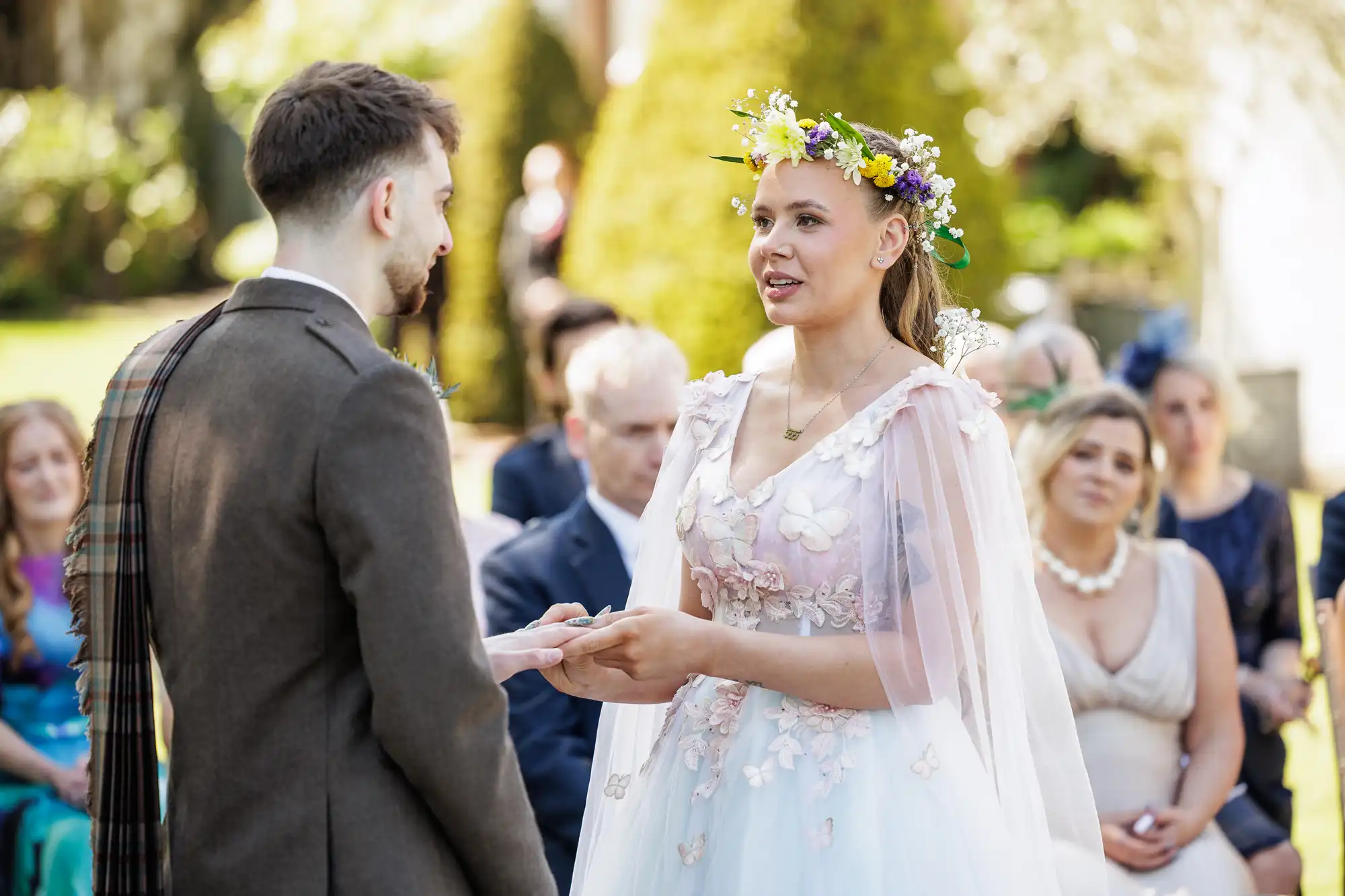 A bride in a flower crown and floral wedding dress holds hands with the groom outdoors during their ceremony, looking emotional. Guests are seated behind them, watching. Sunlight filters through trees, creating a soft, warm atmosphere.