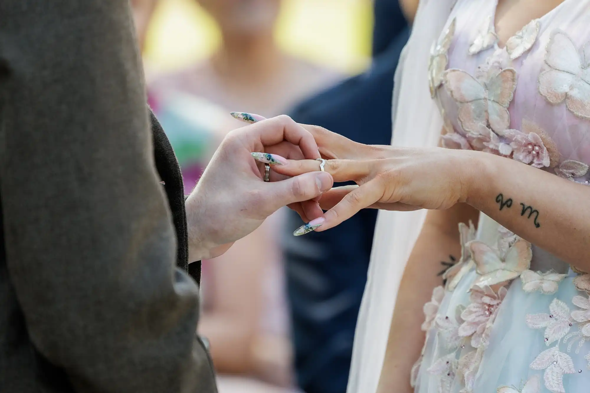 Close-up of a couple exchanging rings at a wedding. The bride wears a pastel floral dress and veil, with long decorated nails and a tattoo on her wrist. The groom wears a dark suit. Their hands are in focus, with blurred guests in the background.