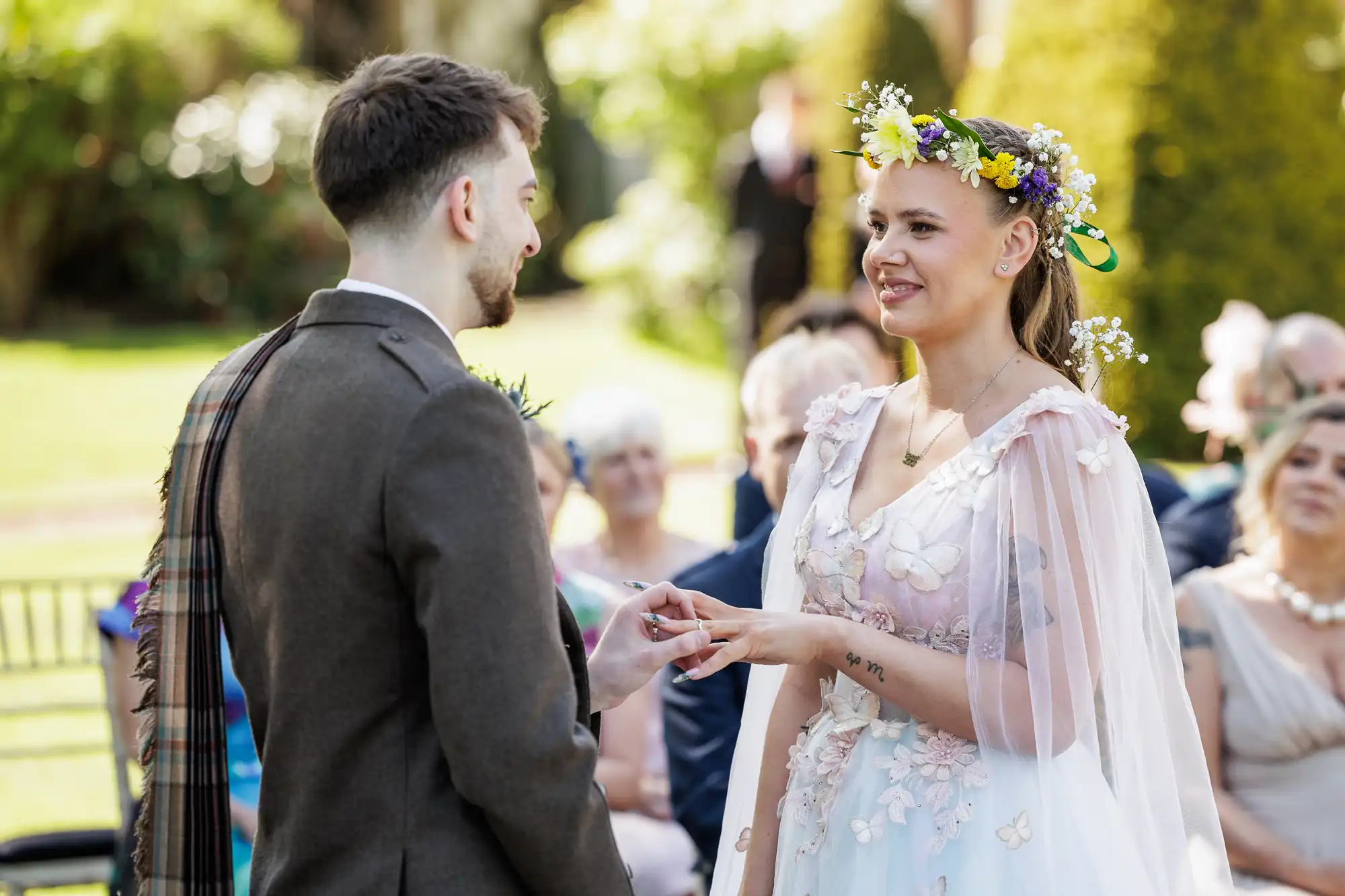 A bride wearing a floral crown and white dress places a ring on the groom’s finger during an outdoor wedding ceremony. Guests watch in the background, seated on chairs, and the setting is bright with sunlight and greenery.