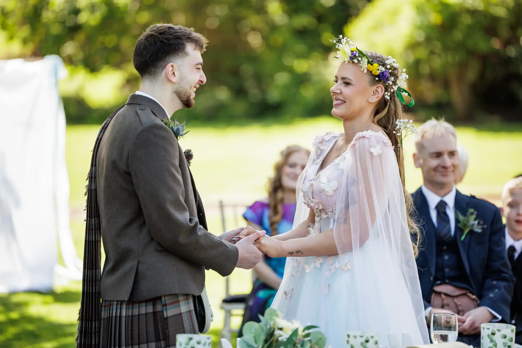 A bride in a floral crown and white gown smiles, holding hands with a groom in a dark waistcoat and kilt. They stand outdoors at a wedding ceremony, with guests seated behind them and lush greenery in the background.