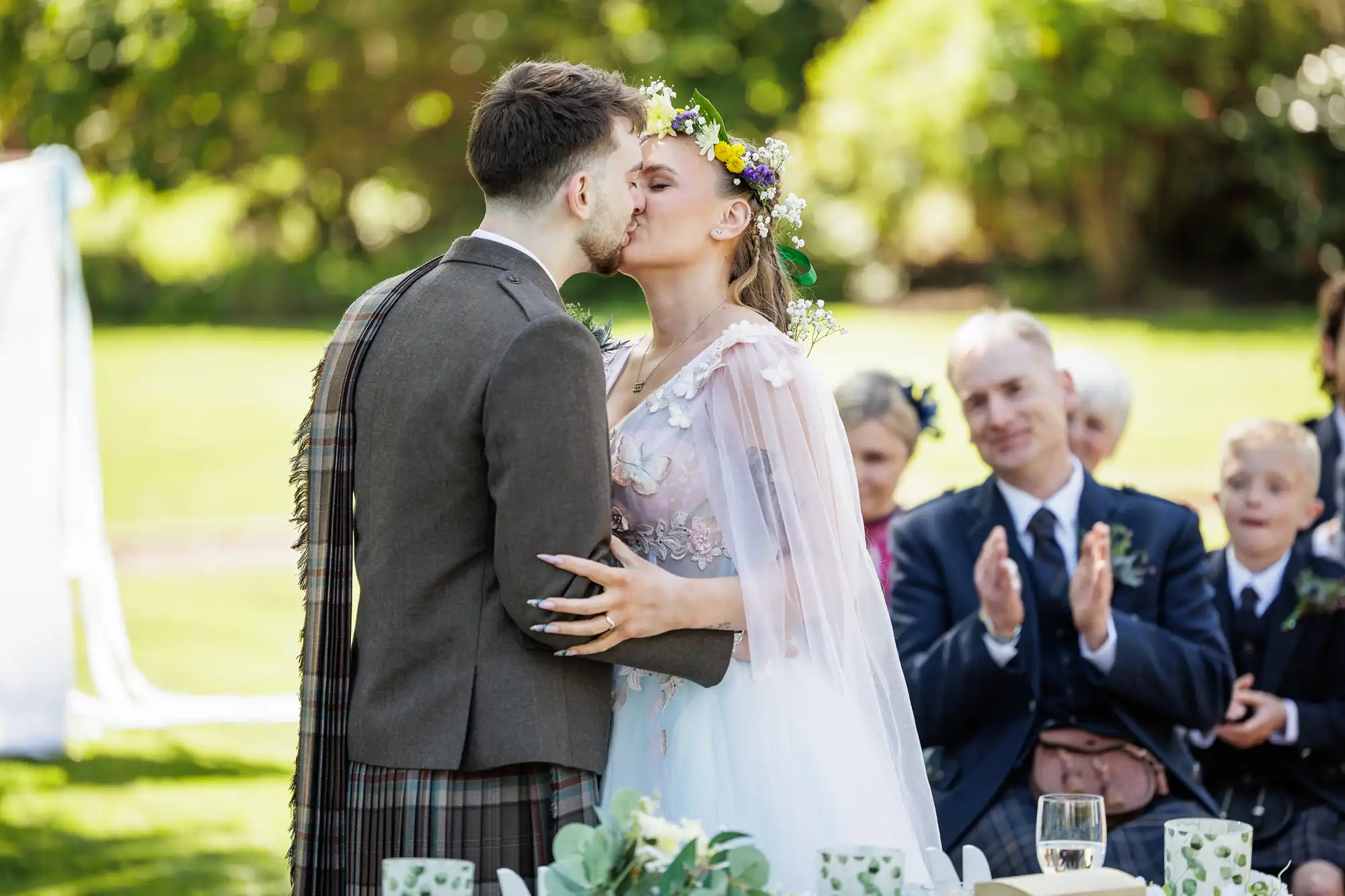 A bride and groom share a kiss outdoors during their wedding. The bride wears a flower crown and white gown, whilst the groom wears a brown jacket with a tartan sash. Guests in formal attire sit and applaud in the blurred background, surrounded by greenery.