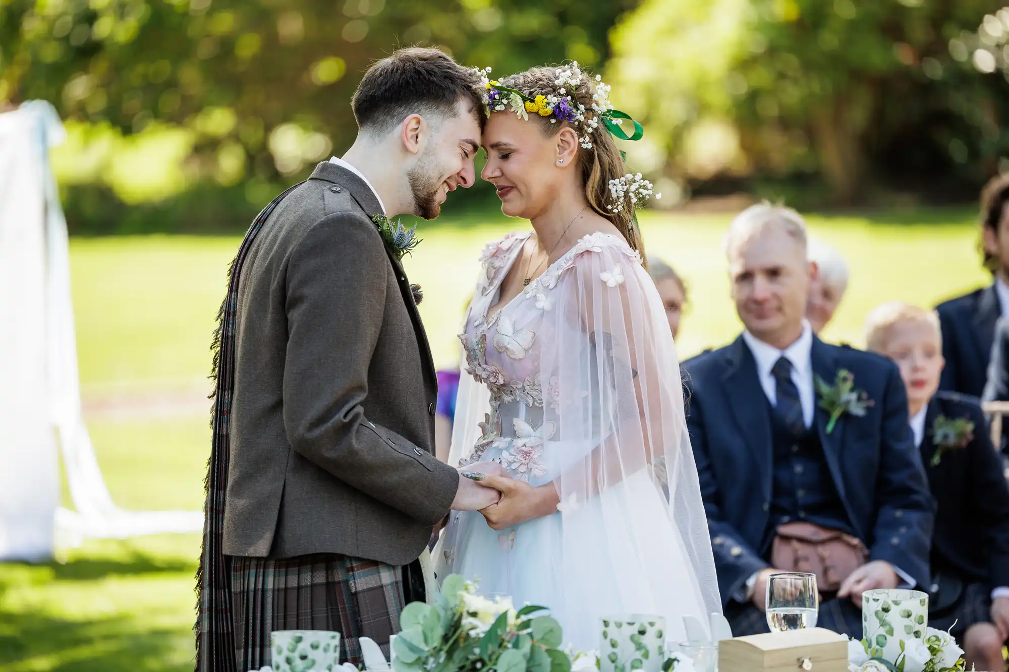 A bride and groom stand close, holding hands and smiling at each other during an outdoor wedding ceremony. The bride wears a floral crown and flowing white dress; the groom wears a brown jacket with a tartan kilt. Guests sit behind them on a sunny day.