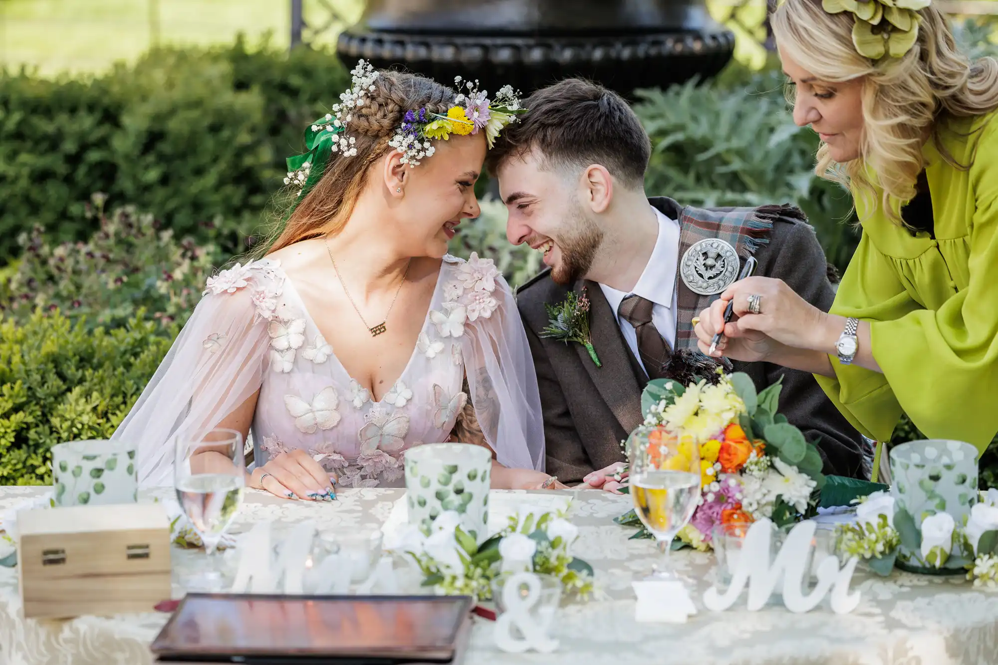 A bride and groom sit closely, smiling at each other, holding hands at an outdoor wedding table decorated with flowers and "Mr & Mrs" signs. A woman in a green dress arranges a bouquet, while greenery and elegant table settings surround them.