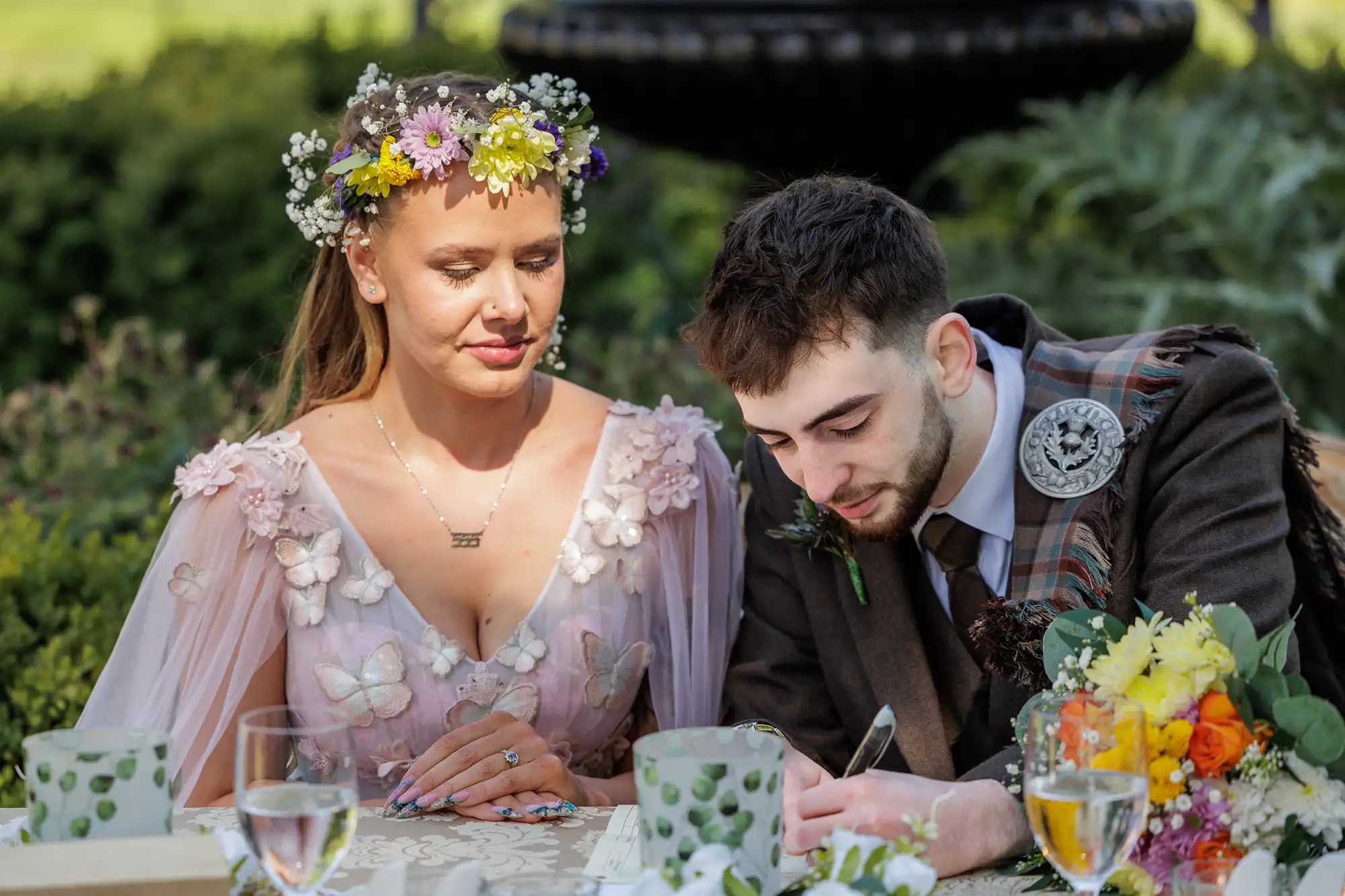 A woman in a floral crown and pink dress with flowers sits beside a man in a dark suit with a sash and brooch. He is writing on paper at an outdoor table decorated with flowers, cups, and glasses, surrounded by greenery and sunlight.