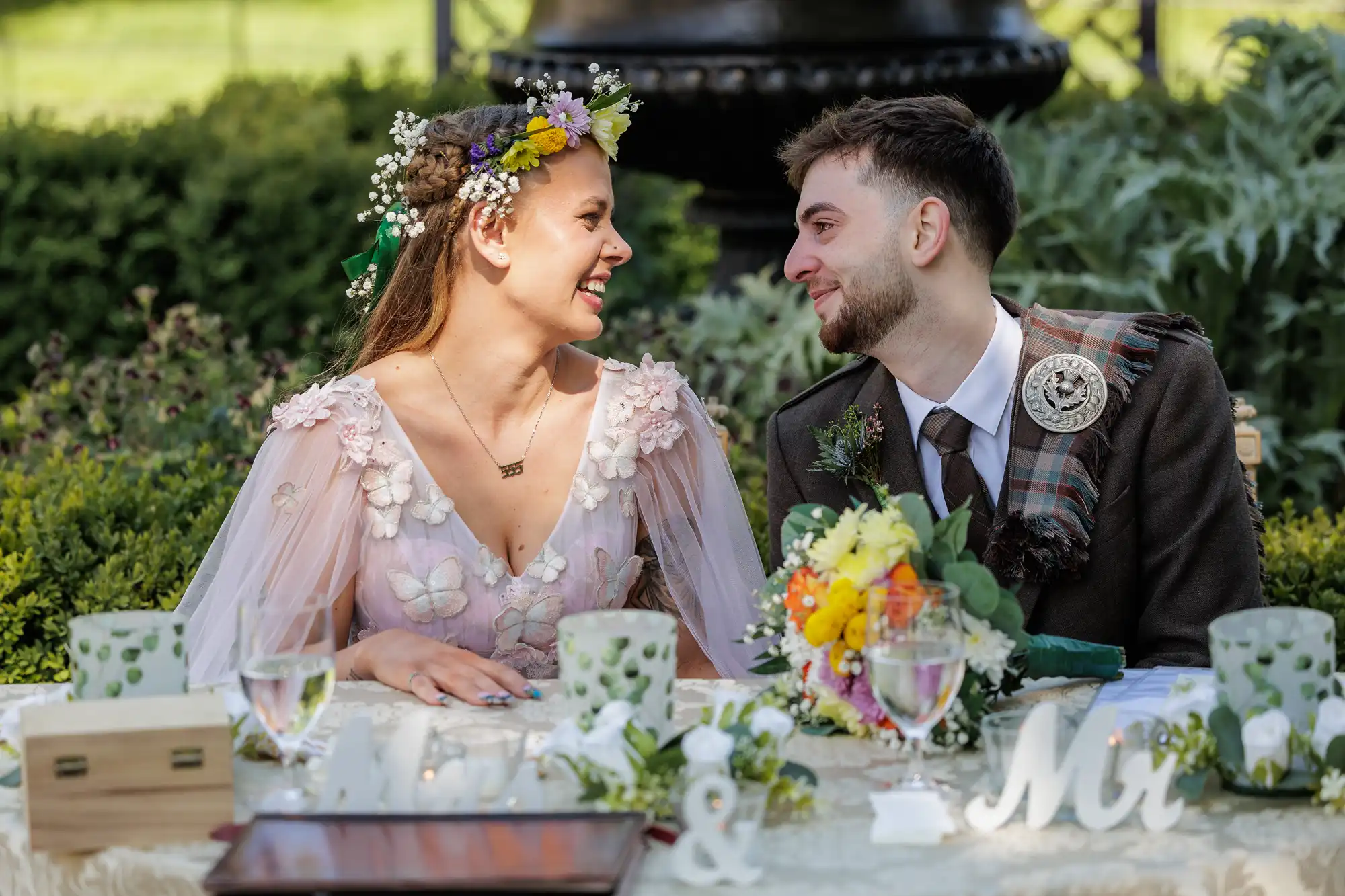 A bride in a floral dress and flower crown smiles at a groom in a suit with a tartan sash. They sit at a decorated table outdoors with “Mr & Mrs” signs, floral arrangements, and green foliage in the background. Both look happy and in love.