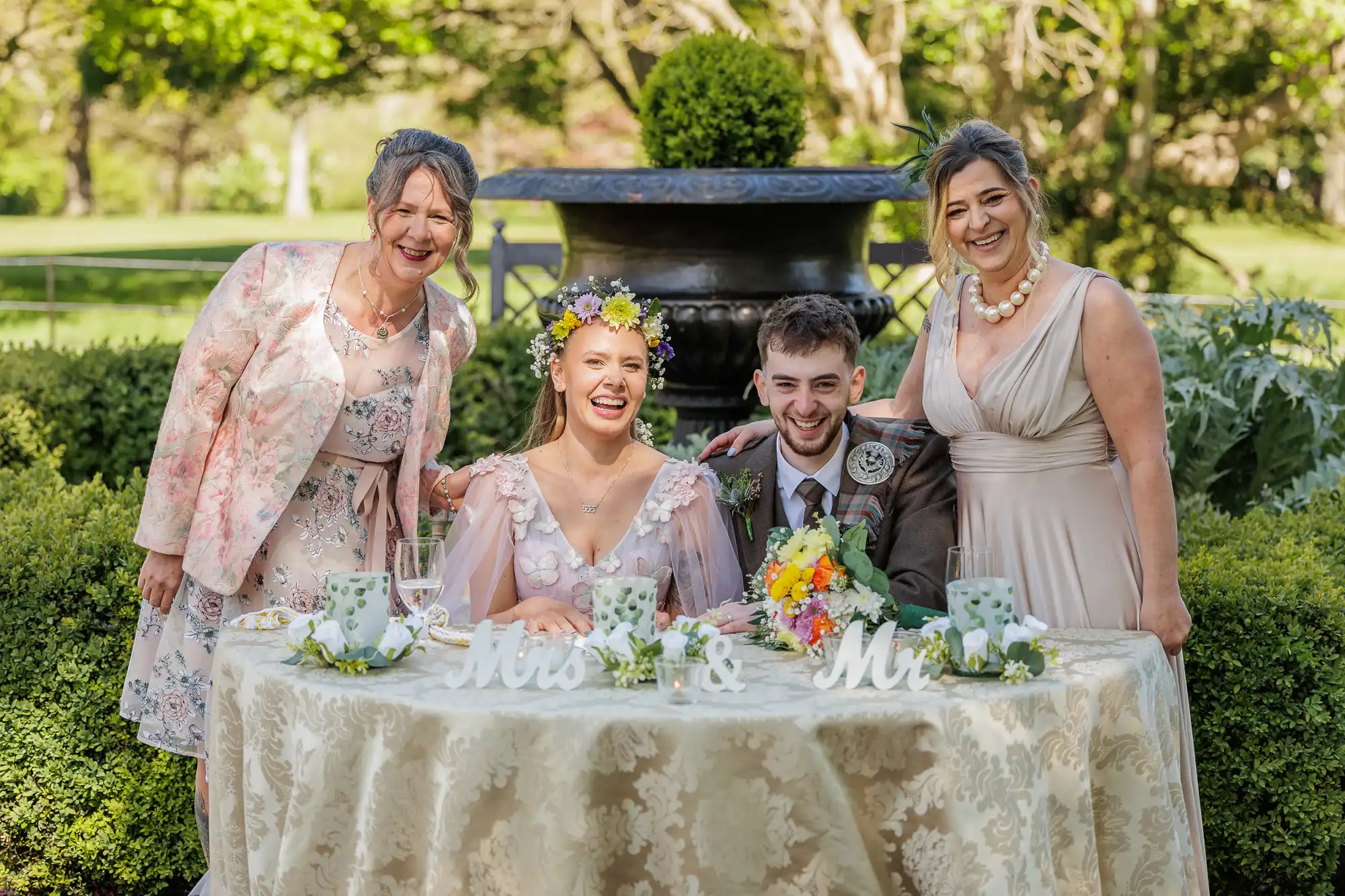 A bride and groom sit at a decorated table outdoors, smiling, with "Mrs & Mr" signs in front. Two women stand behind them, also smiling. The group is dressed in formal attire, surrounded by greenery and sunlight, with a large urn and trees in the background.