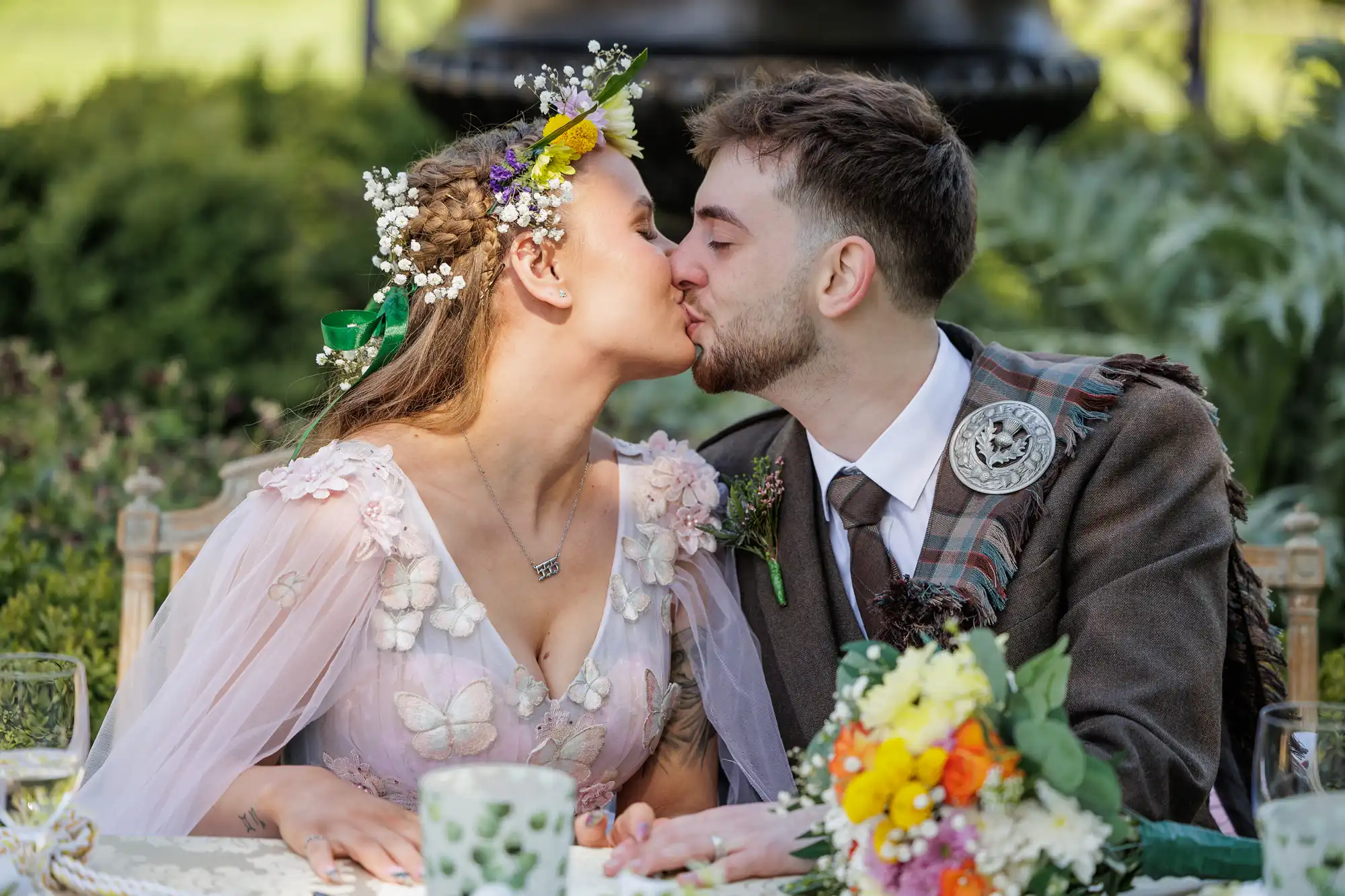 A bride in a floral crown and pastel dress with butterfly details kisses a groom wearing a brown tartan suit and buttonhole at an outdoor table adorned with a bright flower bouquet and greenery in the background.