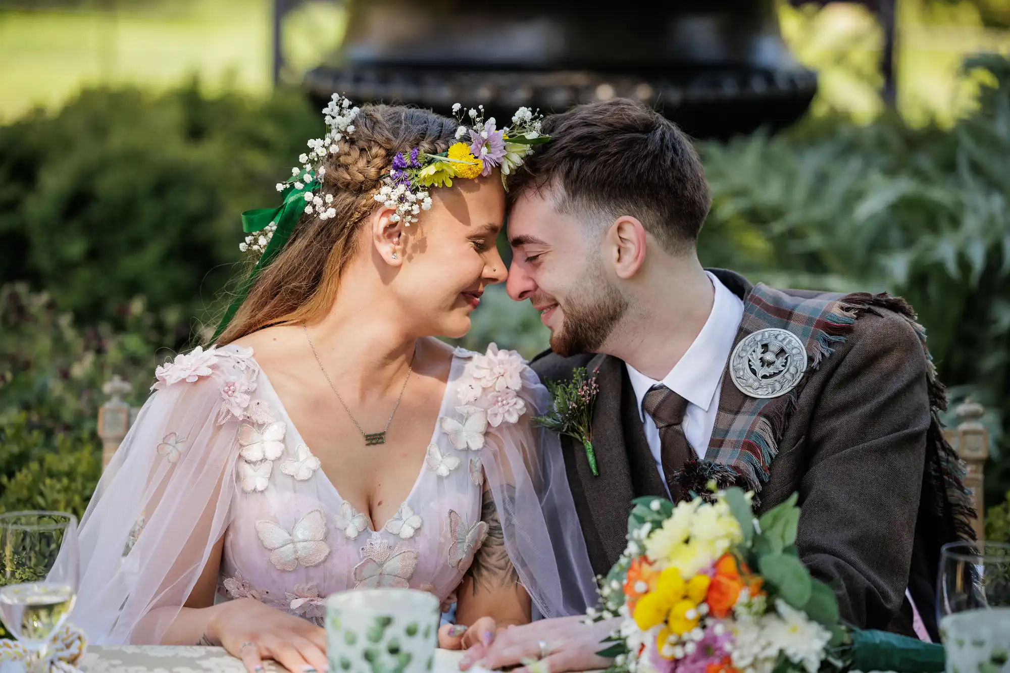 A smiling bride and groom sit close together, touching foreheads. The bride wears a floral gown and a flower crown, while the groom is in a suit with tartan and a decorative brooch. A colourful bouquet rests on the table in front of them. Greenery is in the background.