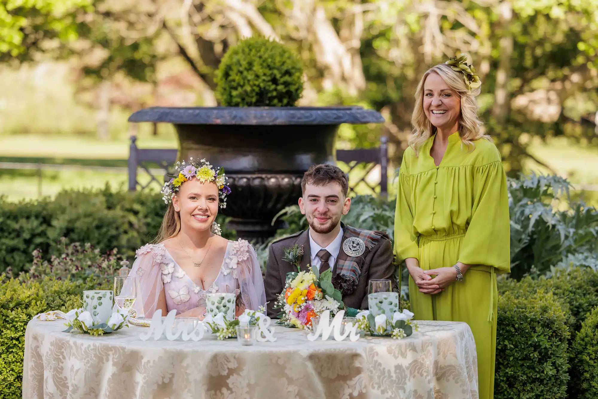 A bride in a floral headpiece and a groom in a kilt sit smiling at a decorated table labelled "Mrs & Mr," with flowers and teacups. Beside them stands a woman in a bright green dress, all outdoors in a lush garden setting.