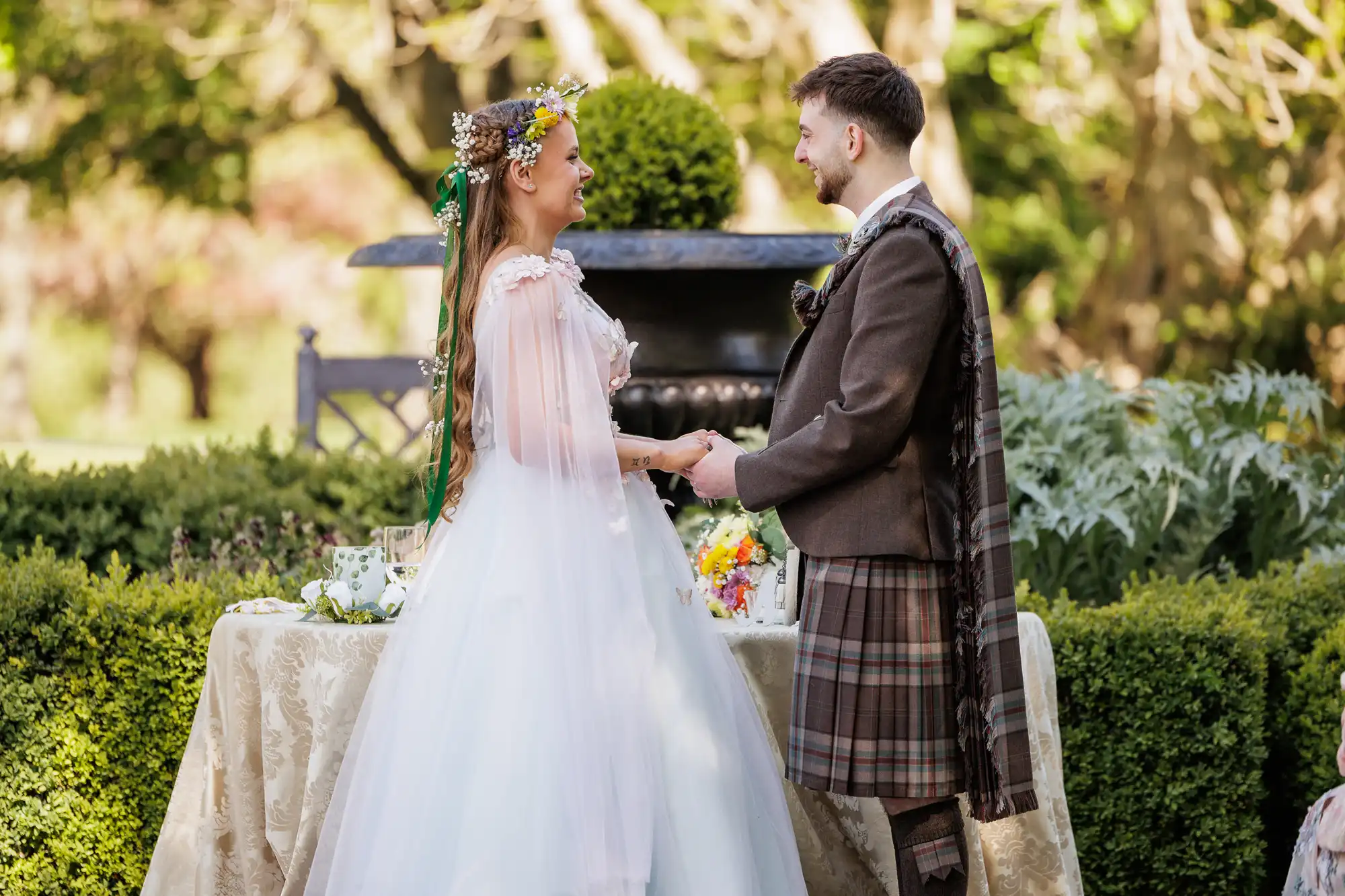 A bride in a white dress with a flower crown and a groom in a brown jacket with a checked kilt stand facing each other, holding hands, outdoors by a table decorated with flowers and a beige cloth, surrounded by greenery and trees.