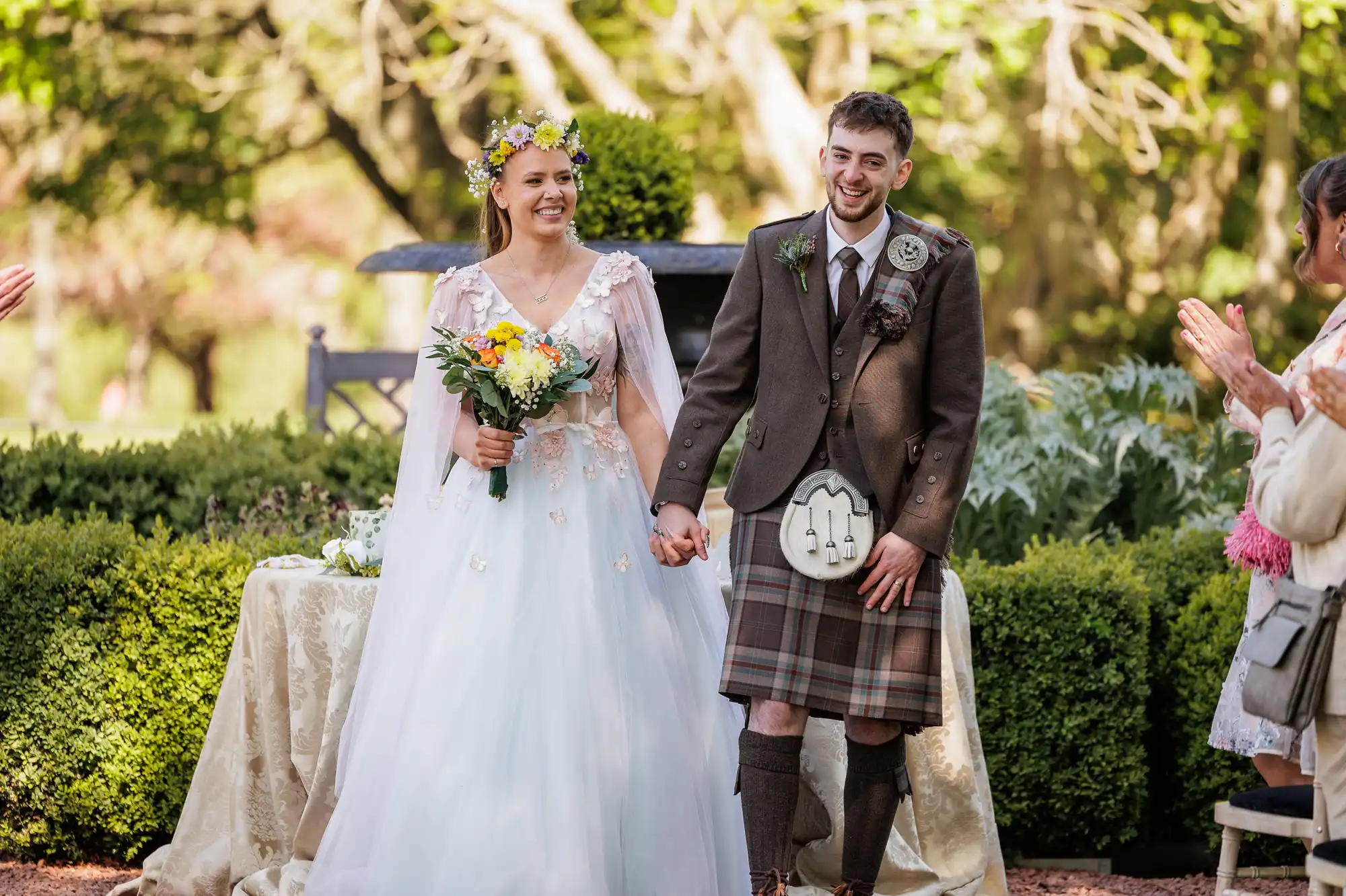 A smiling bride in a white gown and flower crown holds a bouquet, standing hand in hand with a groom in a brown checked kilt outfit. They are outdoors, surrounded by greenery, with people clapping nearby, suggesting a joyful wedding ceremony.