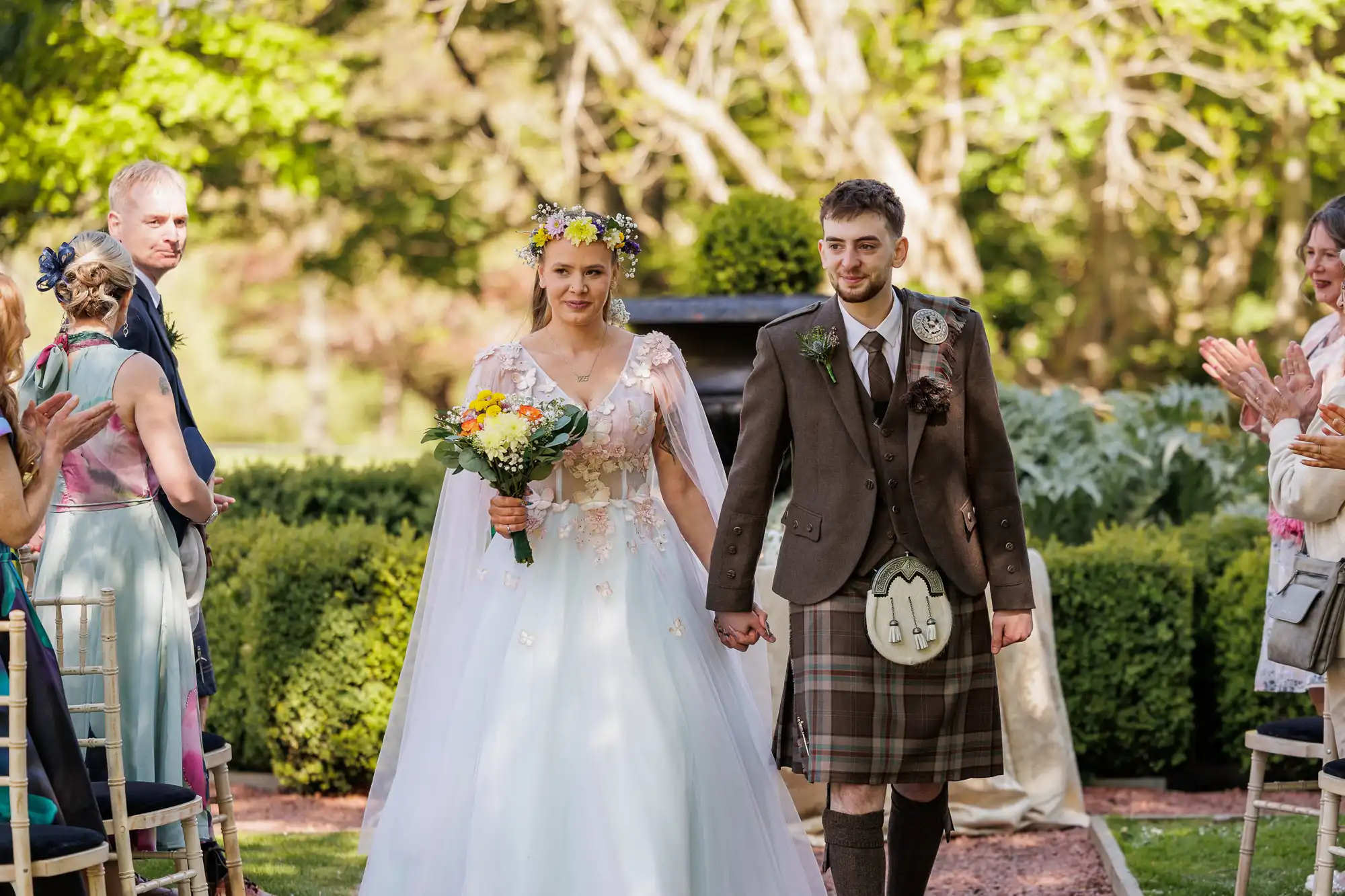 A bride in a floral crown and white dress holds a bouquet and walks hand-in-hand with a groom in a brown jacket, kilt, and sporran. Guests seated on either side clap as the couple walks outdoors, surrounded by greenery and sunlight.
