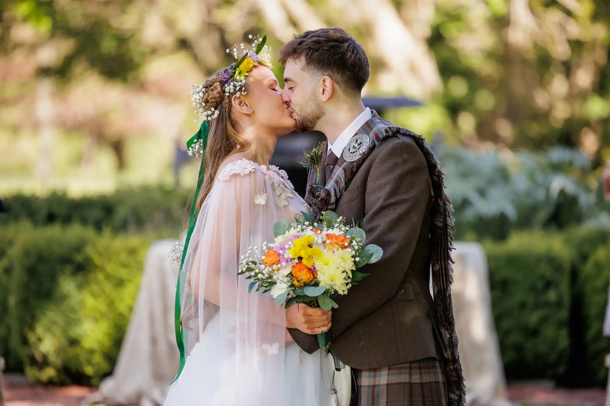 A bride and groom kiss outdoors. The bride wears a white dress, floral crown, and holds a bouquet of yellow, orange, and white flowers. The groom wears a brown jacket and checked kilt. Greenery and blurred trees fill the background.
