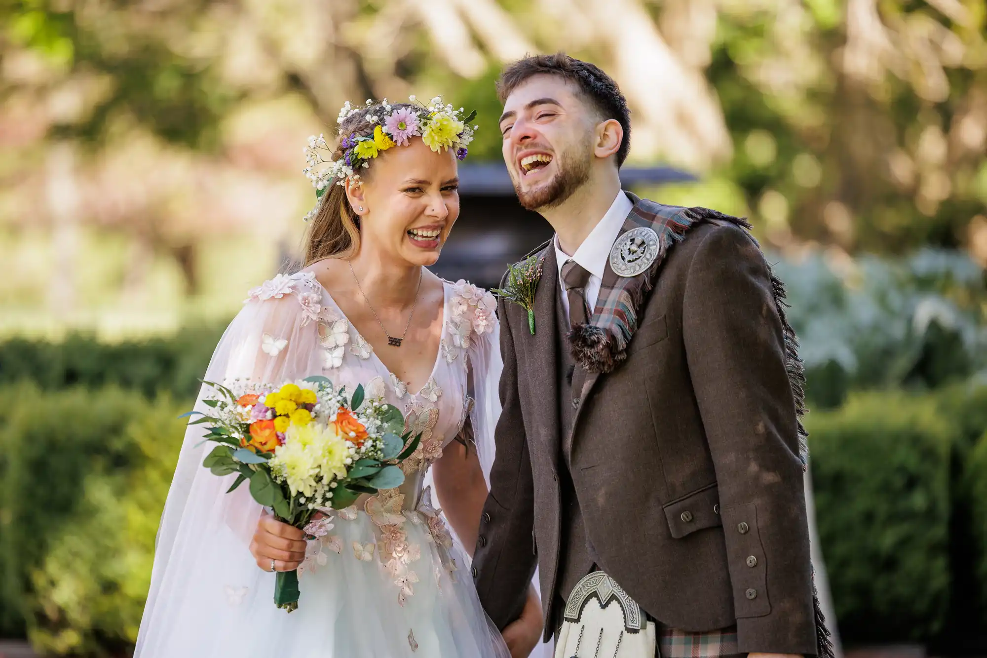 A joyful bride in a floral crown and pastel dress holding a bouquet stands beside a laughing groom in a brown jacket, checked kilt, and sporran. They are outdoors, surrounded by greenery and sunlight, appearing happy and relaxed on their wedding day.