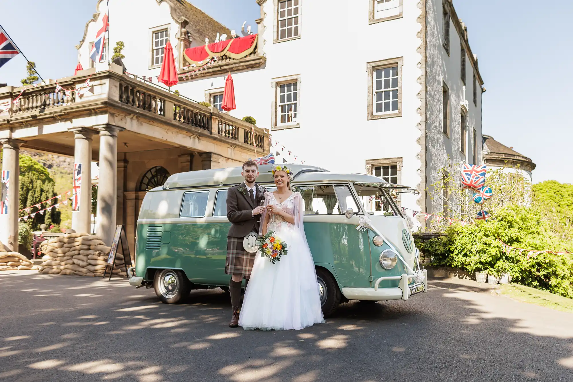 A bride in a white gown and a groom in a kilt stand arm-in-arm beside a vintage green van, smiling. They are outside a grand building decorated with British flags and bunting, with a clear blue sky overhead.