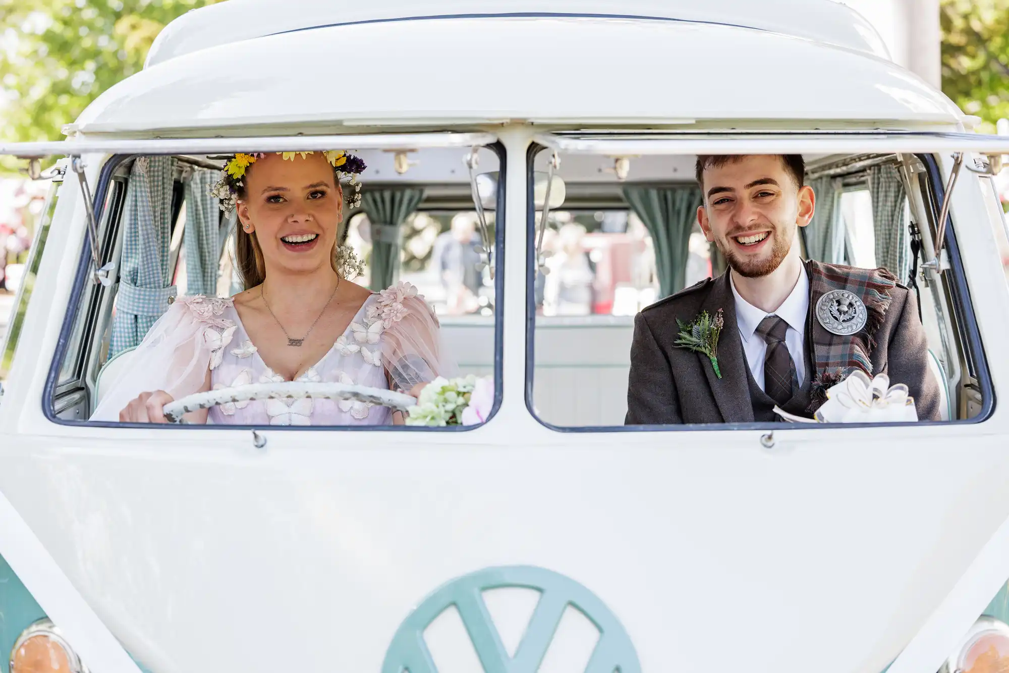A smiling bride in a floral crown and pastel dress sits at the wheel of a vintage VW van, next to a happy groom in a checked suit with buttonhole. They look joyful, surrounded by sunlight and greenery outside the vehicle.
