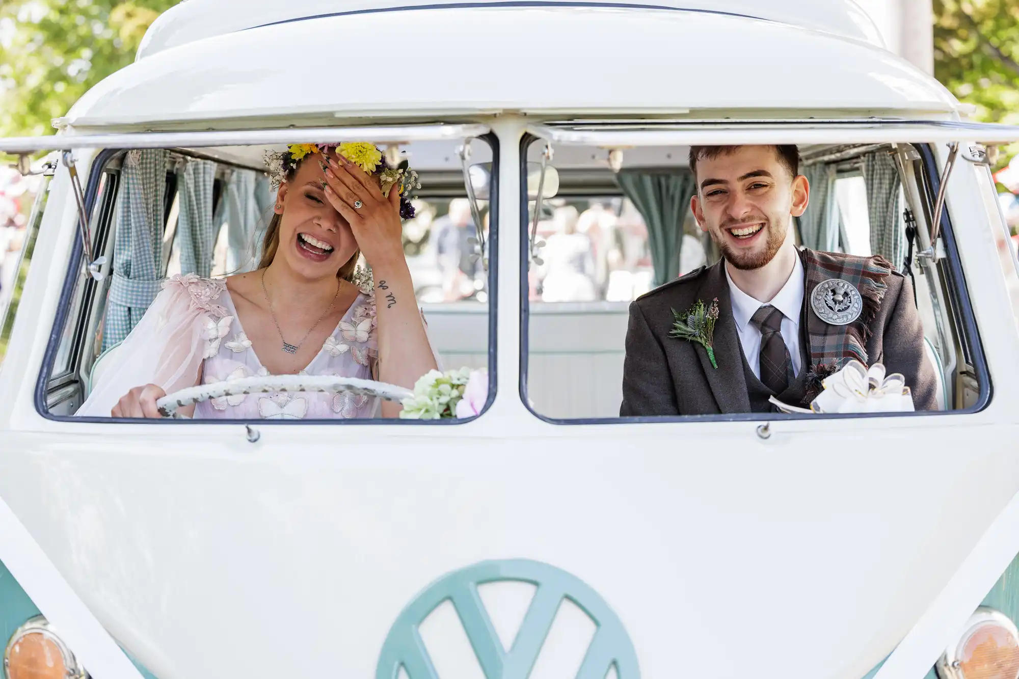 A bride and groom sit in the front seats of a vintage white van. The bride, wearing a flower crown and light dress, laughs with her hand covering part of her face. The groom in a suit and kilt smiles happily beside her. Both seem joyful and excited.