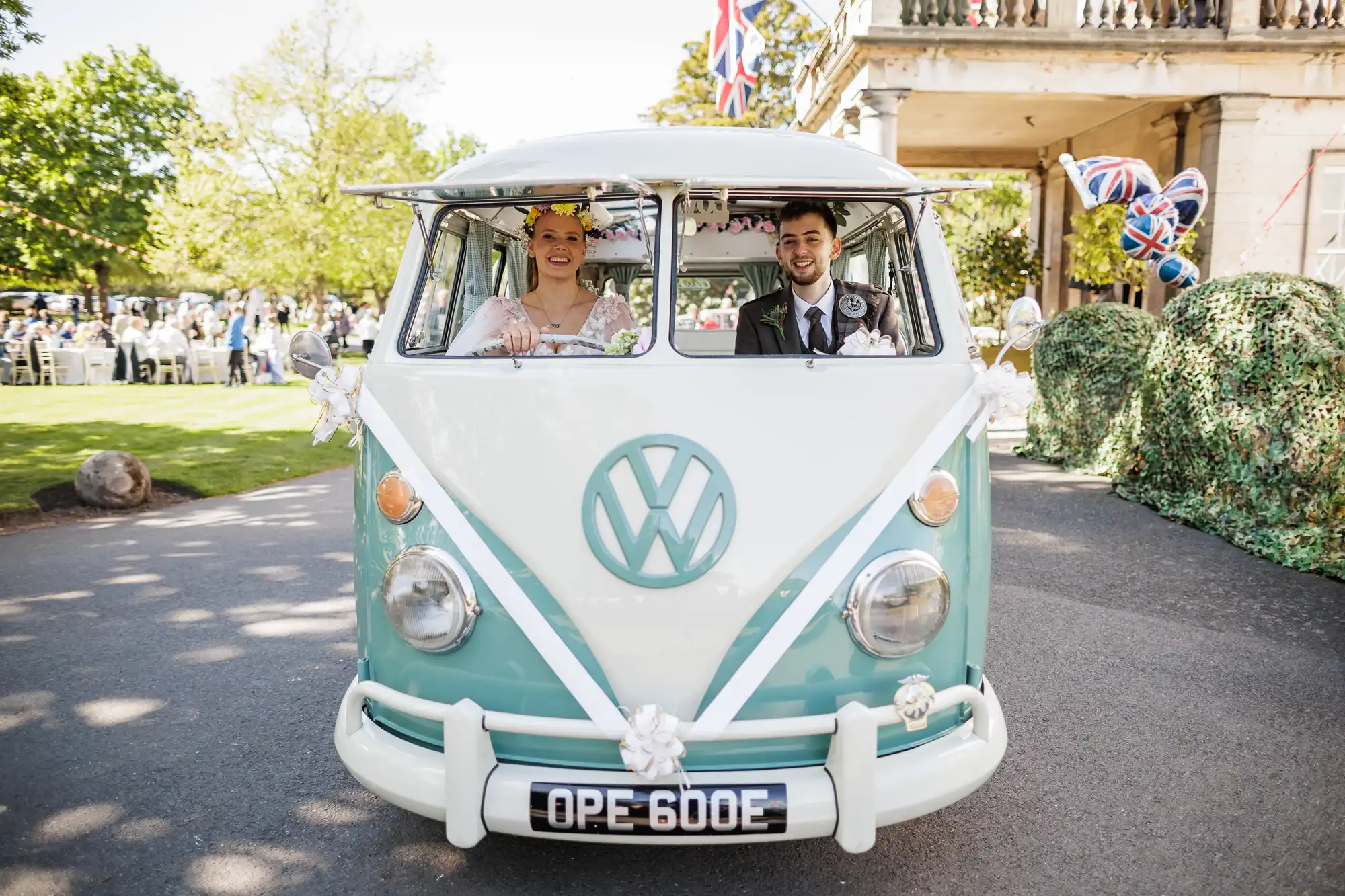 A bride and groom smile from inside a vintage, pale blue and white Volkswagen van decorated with white ribbons and flowers, parked outdoors near a crowd, greenery, and British flags, suggesting a festive wedding celebration.