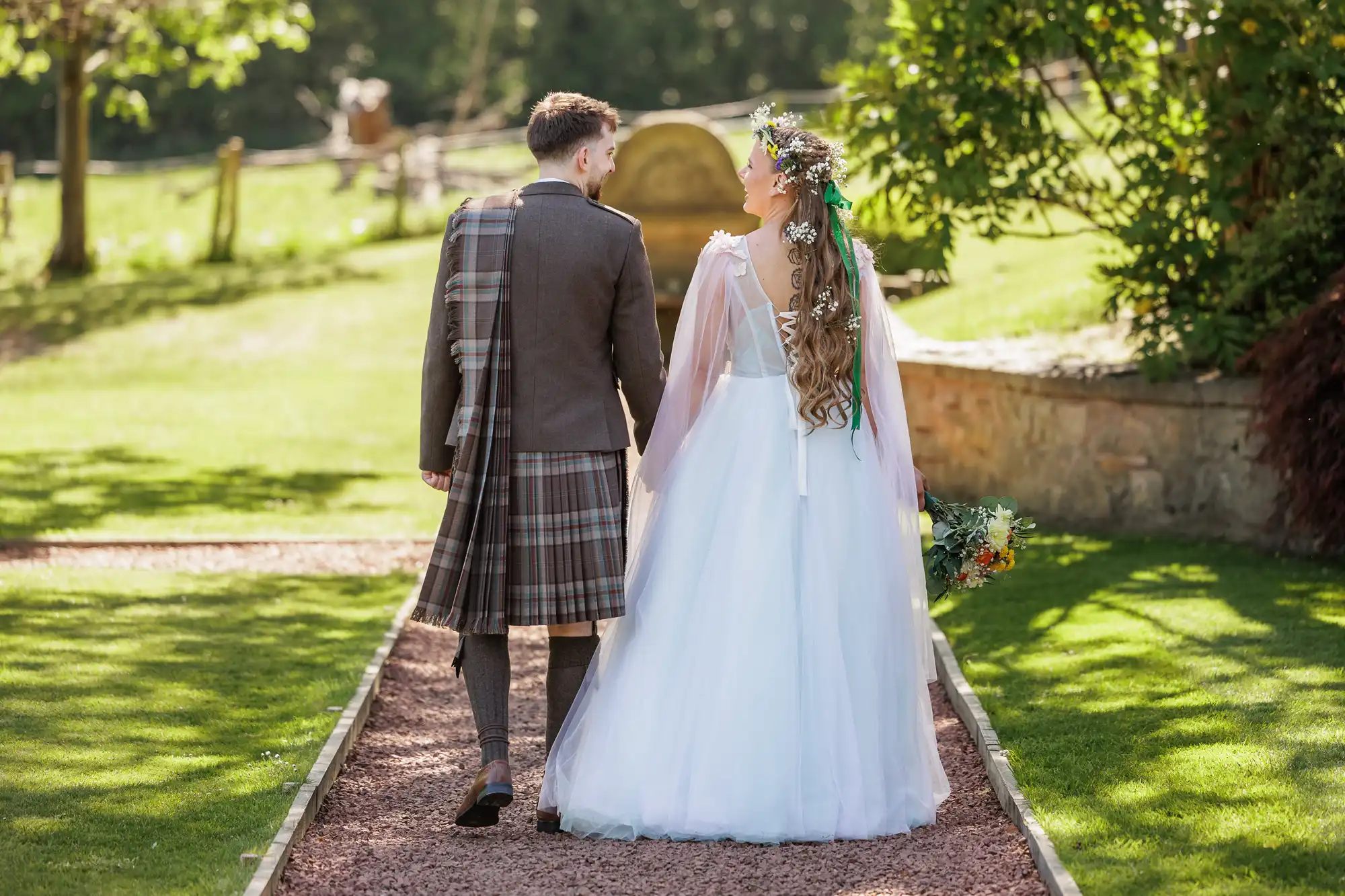 A bride in a white dress with a floral crown and a groom in a brown checked kilt walk hand-in-hand down a garden path, outdoors on a sunny day, with green grass, trees, and a stone wall in the background.