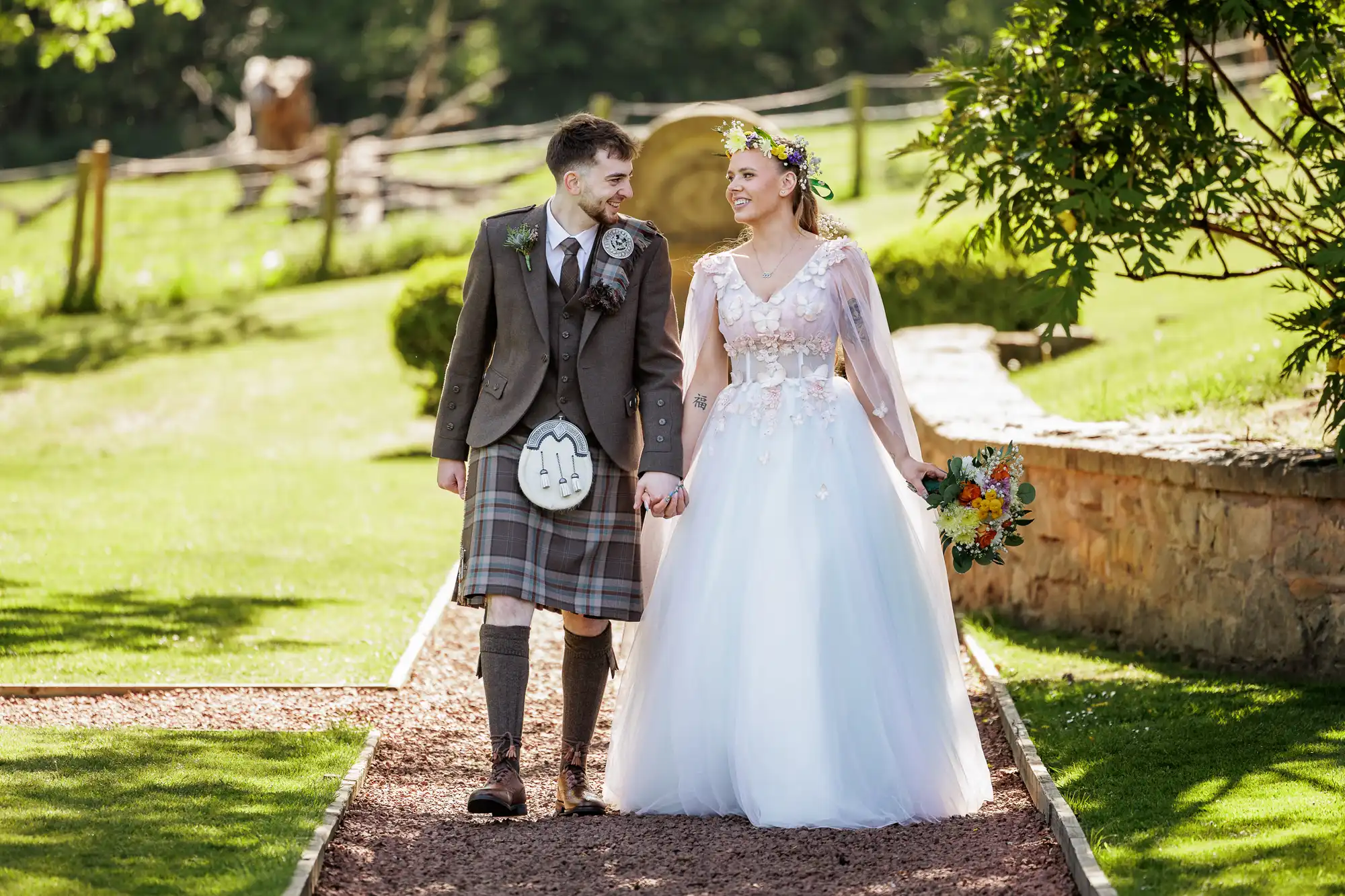 A couple walks hand in hand outdoors. The man wears a brown tweed jacket, a checked kilt, and a sporran; the woman wears a white gown with floral details and a flower crown, holding a bouquet. They smile at each other, surrounded by greenery.