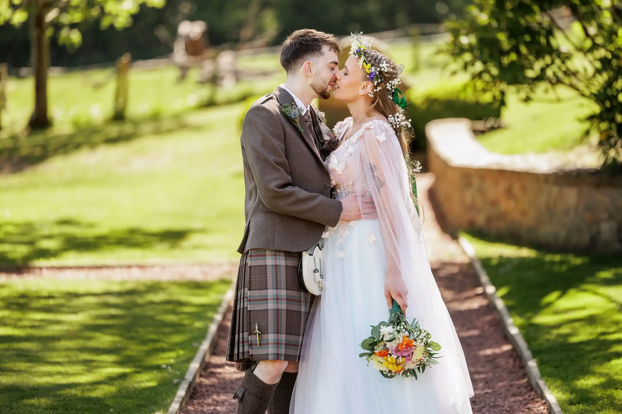 A couple kiss outdoors on a path. The man wears a brown jacket and a traditional kilt; the woman wears a white wedding dress and floral crown, holding a bouquet. Sunlight brightens the lush green garden background.