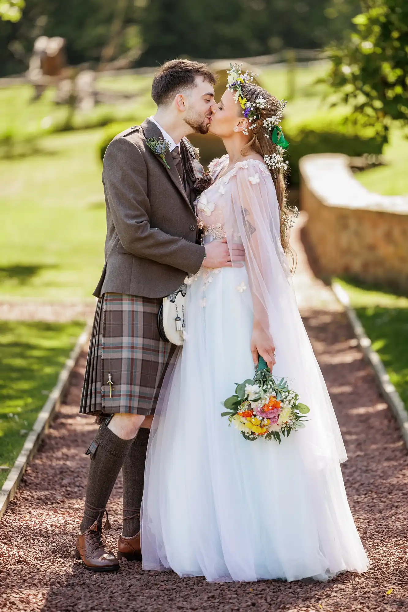 A bride and groom kiss outdoors on a path. The groom wears a brown jacket and tartan kilt with a sporran, while the bride wears a white dress, floral crown, and veil, holding a bouquet of flowers. Green grass and trees fill the bright background.