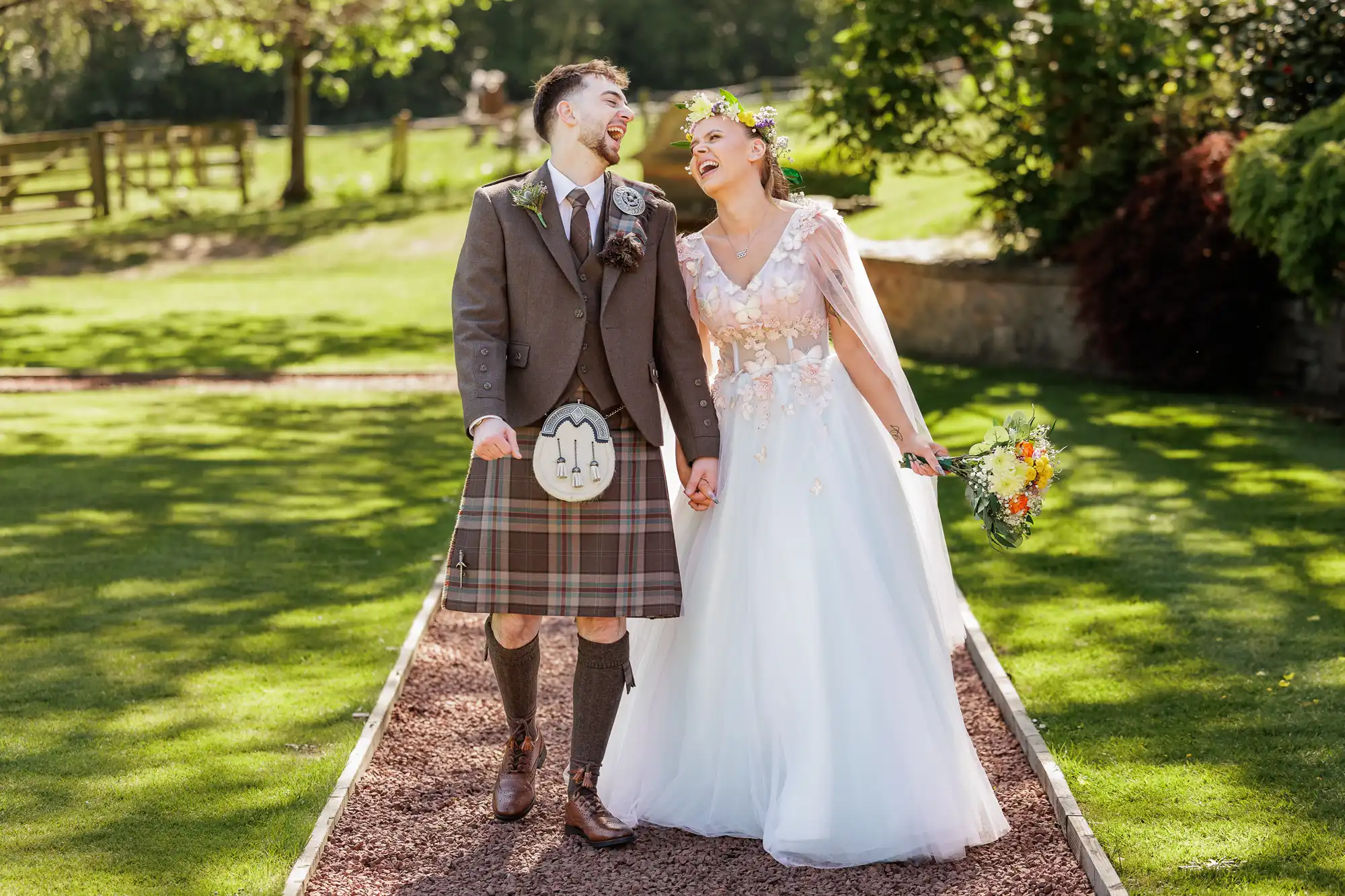 A joyful couple walk hand in hand outdoors. The groom wears a brown jacket, tartan kilt, and sporran; the bride wears a white gown with floral details and a flower crown, holding a bouquet. Both are smiling, surrounded by lush greenery on a sunny day.