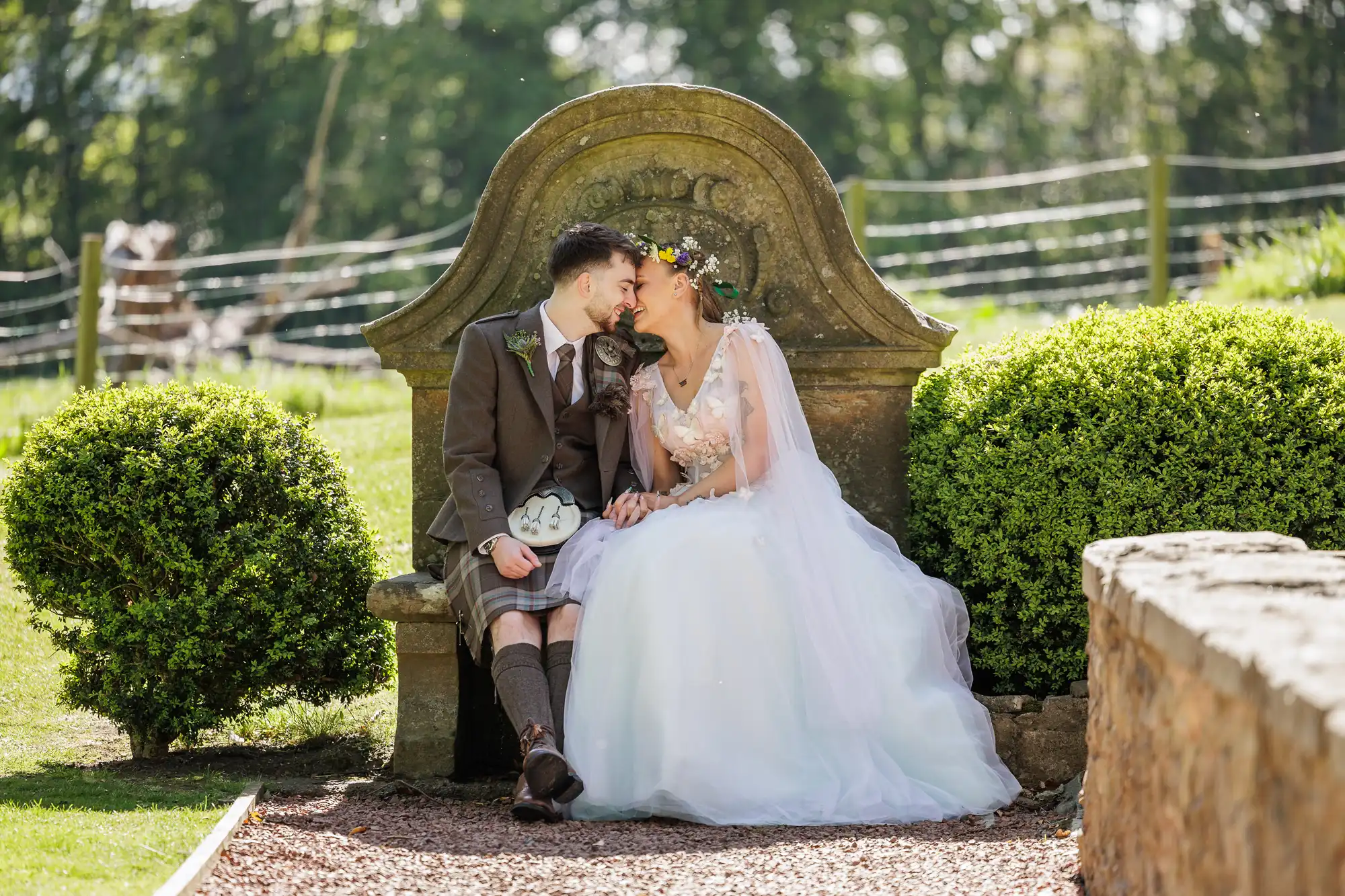 A bride in a white gown and a groom in a kilt sit closely on a stone bench outdoors, touching foreheads and smiling. Green bushes flank the bench; the background features sunlit grass, trees, and a fence, suggesting a countryside wedding setting.