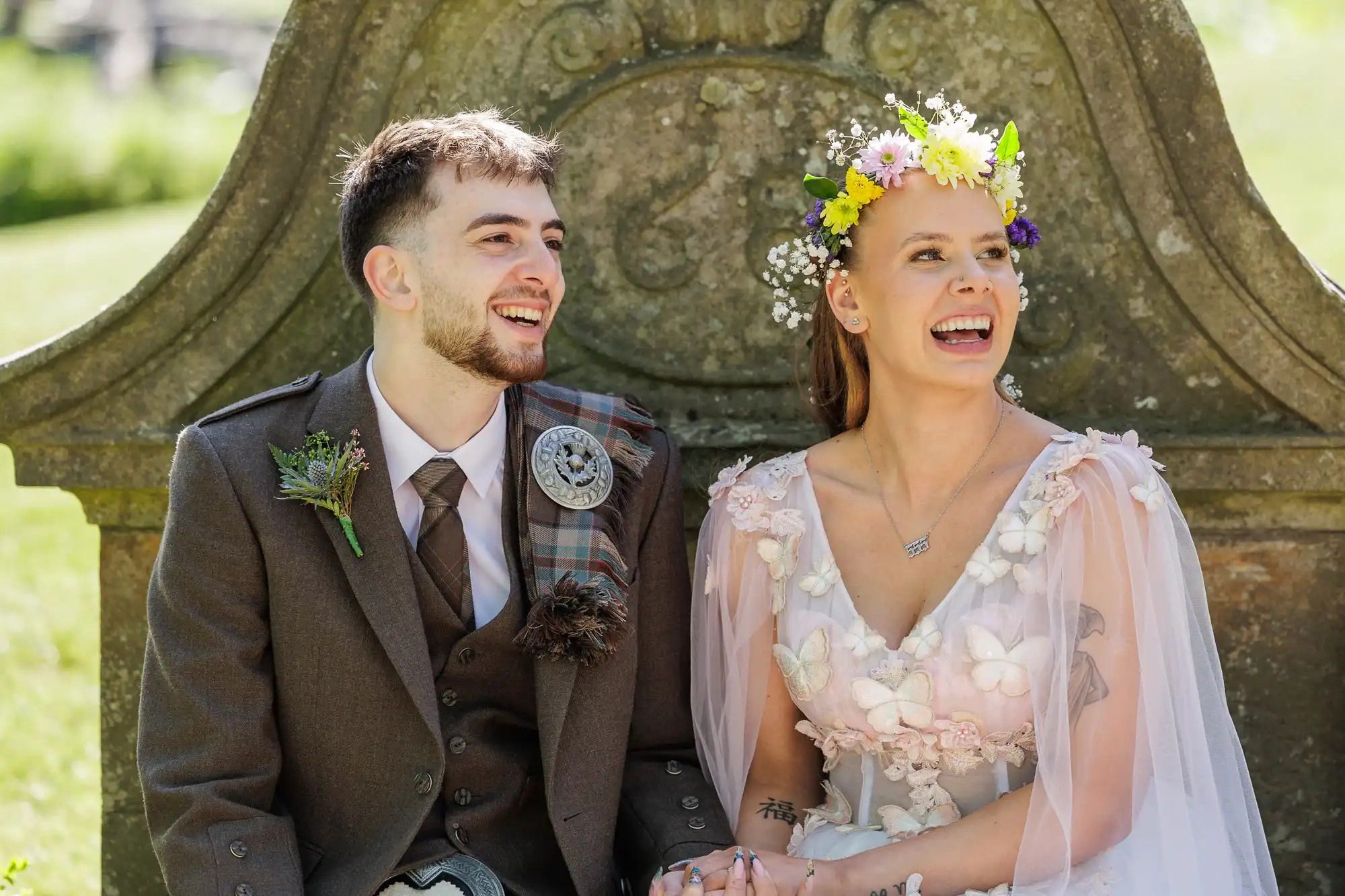 A smiling couple sit outdoors on a stone bench. The man wears a brown suit with a tie and kilt, and the woman wears a floral crown and a pale pink dress adorned with flowers and butterflies. Both look happy, enjoying a sunny day.
