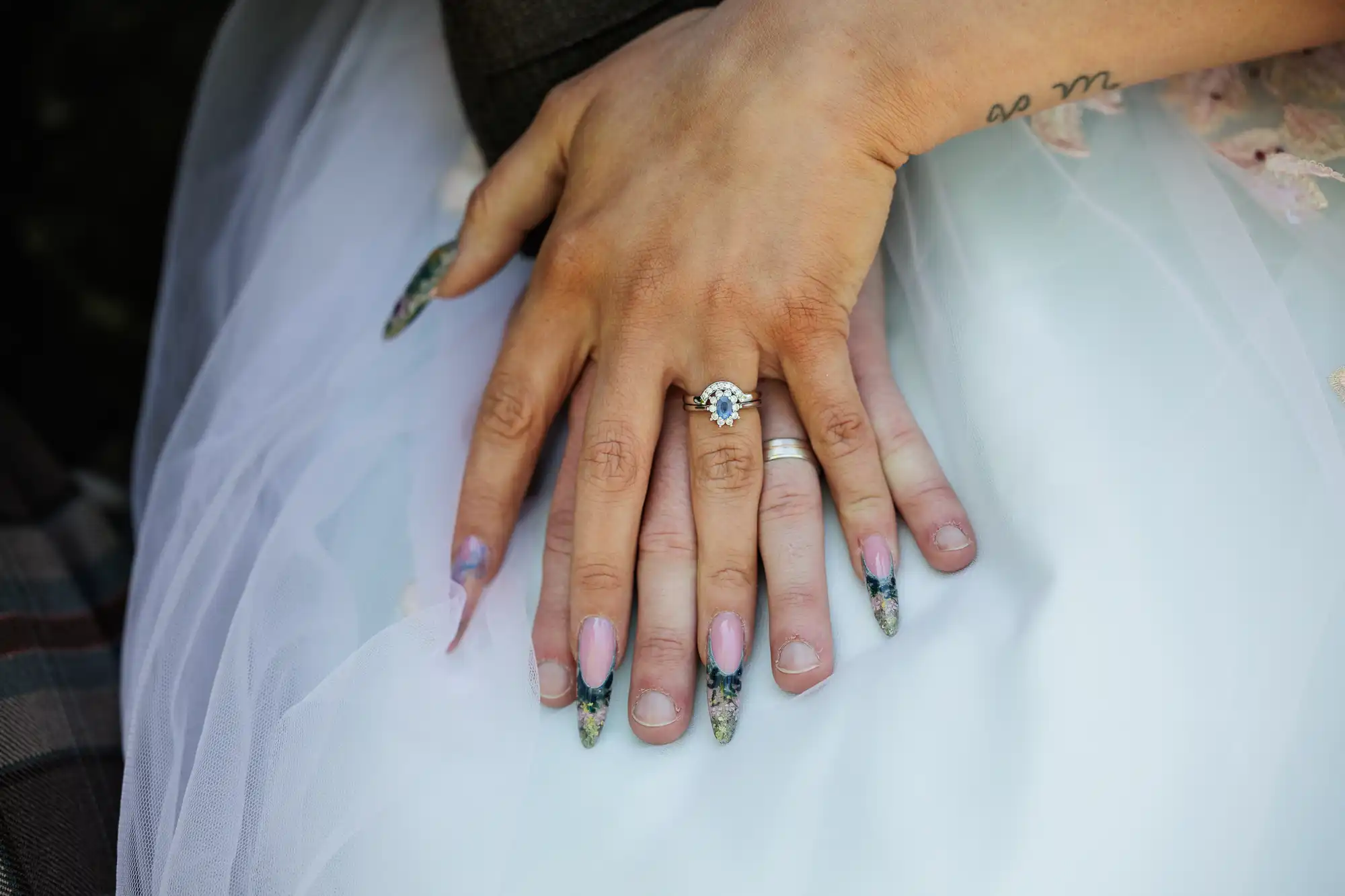 Two hands rest on a white dress: one with long, decorated nails and a blue gemstone engagement ring, the other with a simple silver wedding band. The hand with decorated nails also has a tattoo on the wrist. Both hands are gently touching.