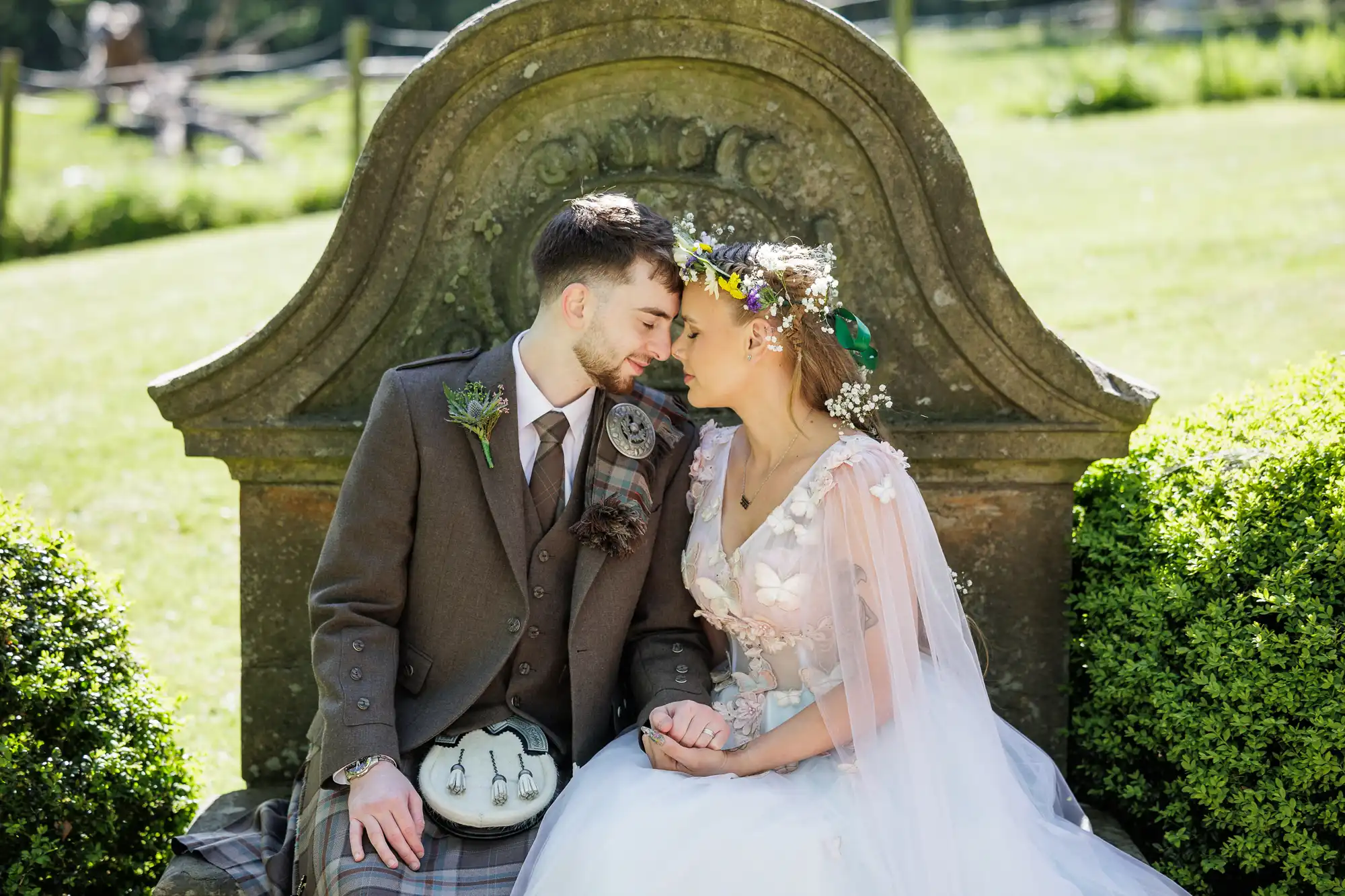 A bride and groom sit close together, touching foreheads and holding hands on a stone bench in a garden. The groom wears a brown checked kilt outfit; the bride wears a white dress with floral details and a flower crown. Both look happy and peaceful.