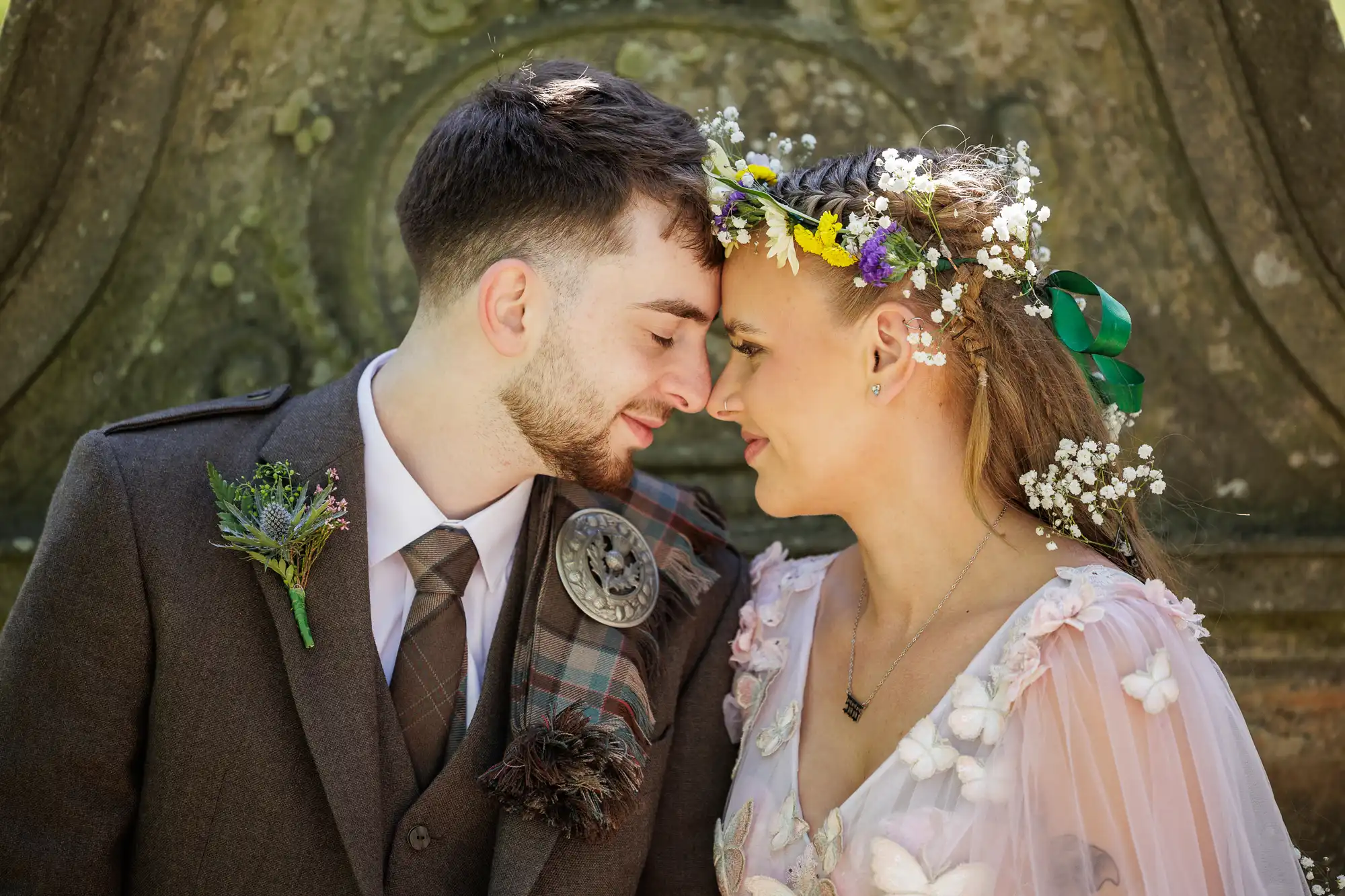 A couple in wedding attire touch foreheads and smile gently. The groom wears a brown suit, tartan sash, and buttonhole, while the bride has a floral crown and pastel dress with flower details. The background is an ornate, mossy stone wall.