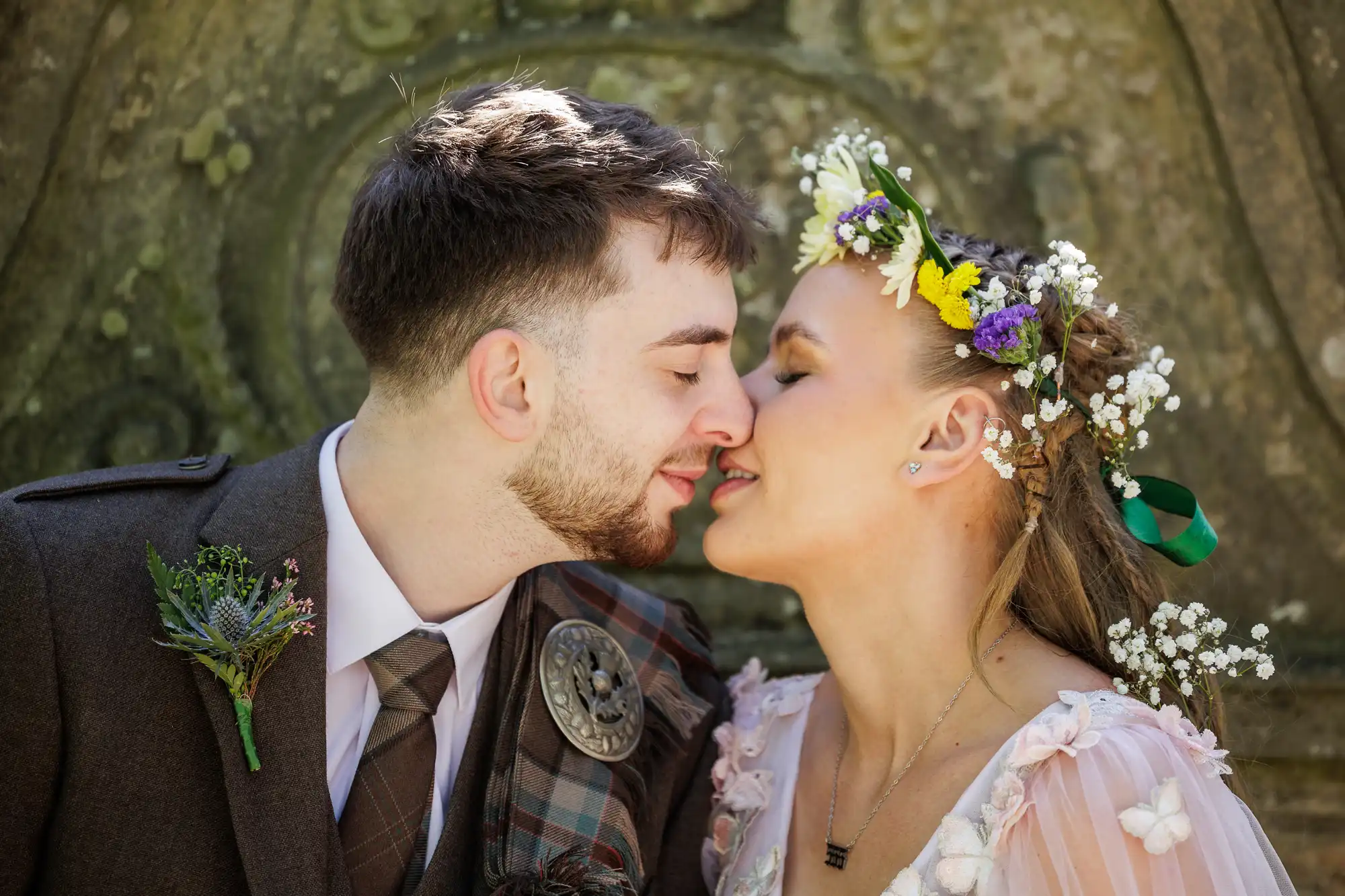 A couple dressed in wedding attire lean in for a kiss. The groom wears a brown suit with a tartan sash and buttonhole, while the bride has a floral crown and light pink dress. Sunlight highlights their faces against a stone background.