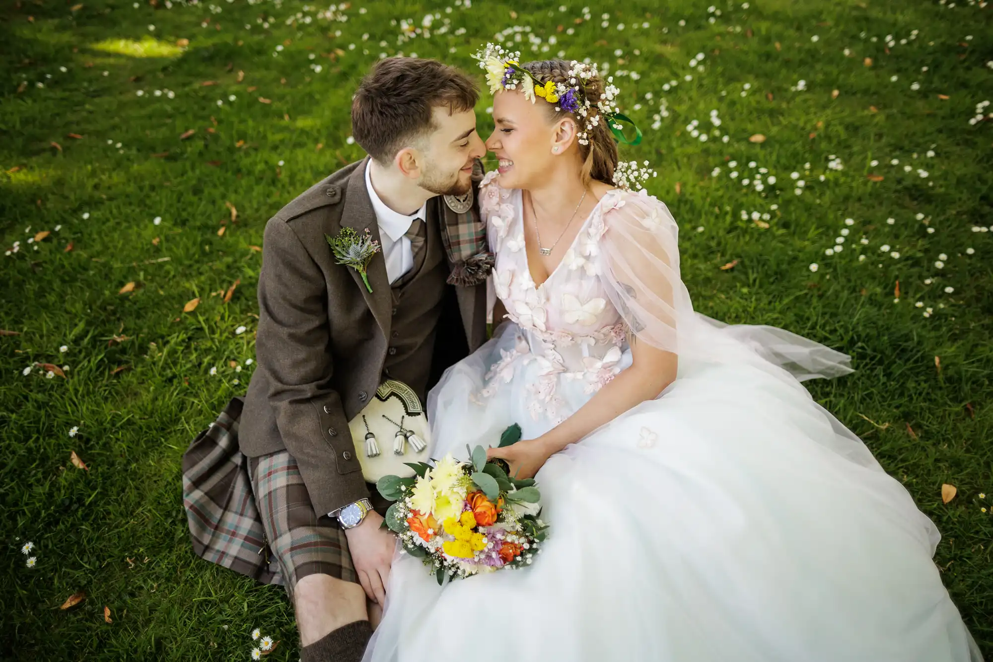 A bride and groom sit on grass, smiling at each other. The bride wears a white gown with floral details and a flower crown, holding a bouquet of colourful flowers. The groom wears a brown jacket, checked kilt, and sporran. White flowers dot the grass around them.
