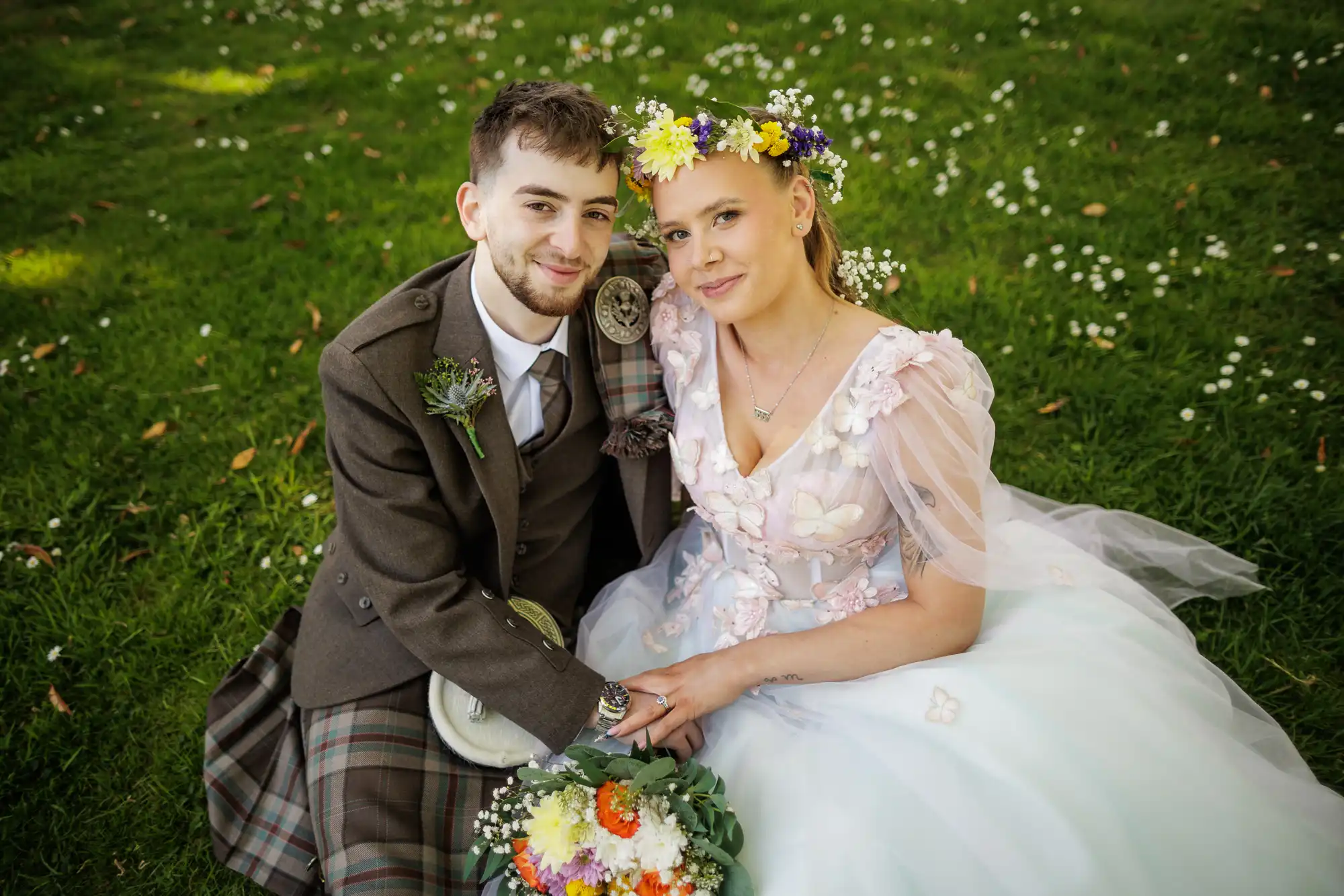 A couple sit on grass, dressed in wedding attire. The groom wears a brown kilt and jacket; the bride wears a light, floral gown with a colourful flower crown. They hold hands and smile gently at the camera. A bouquet of flowers rests in the bride’s lap.