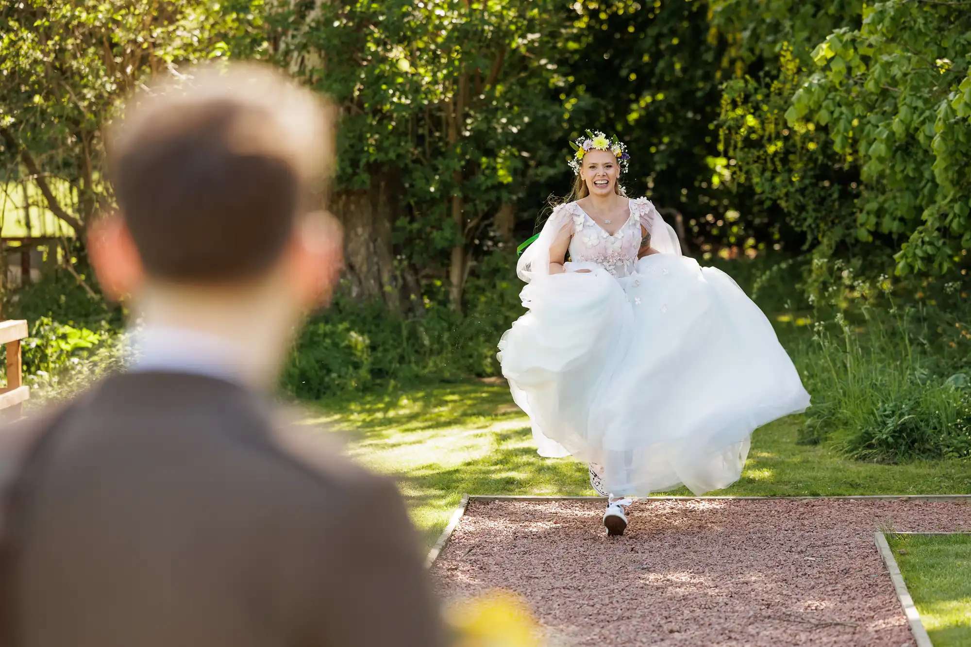 A bride in a white dress and floral crown joyfully runs towards a person in a suit, who is blurred in the foreground. They are outdoors on a sunny day, surrounded by lush green trees and grass. The bride is smiling and holding up her dress.