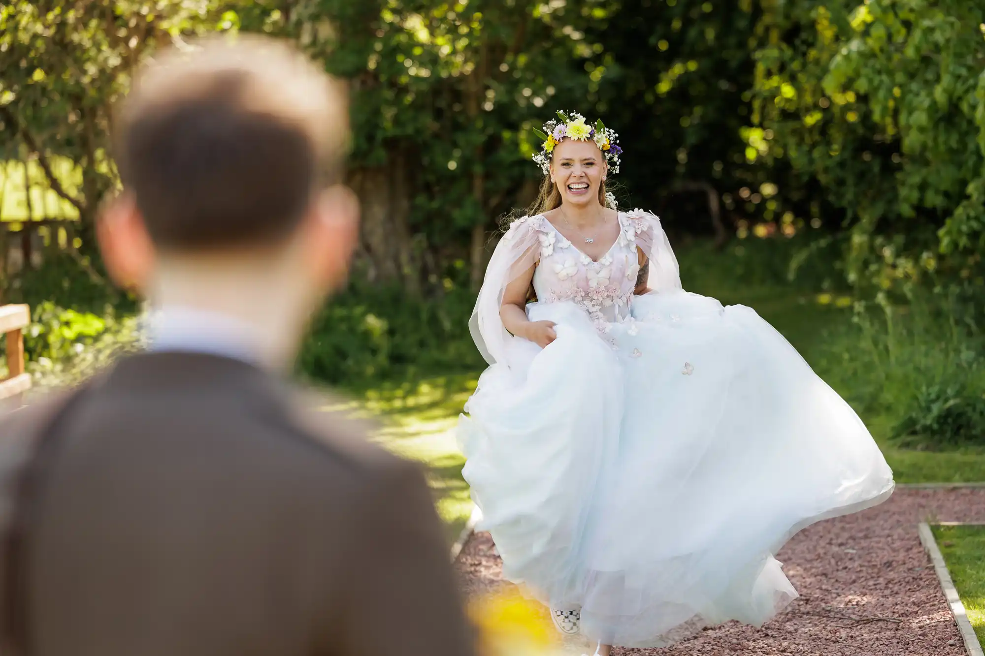 A smiling bride in a white dress and flower crown lifts her dress slightly as she approaches a person in a suit, whose back is to the camera, outdoors in a sunlit, green garden setting.