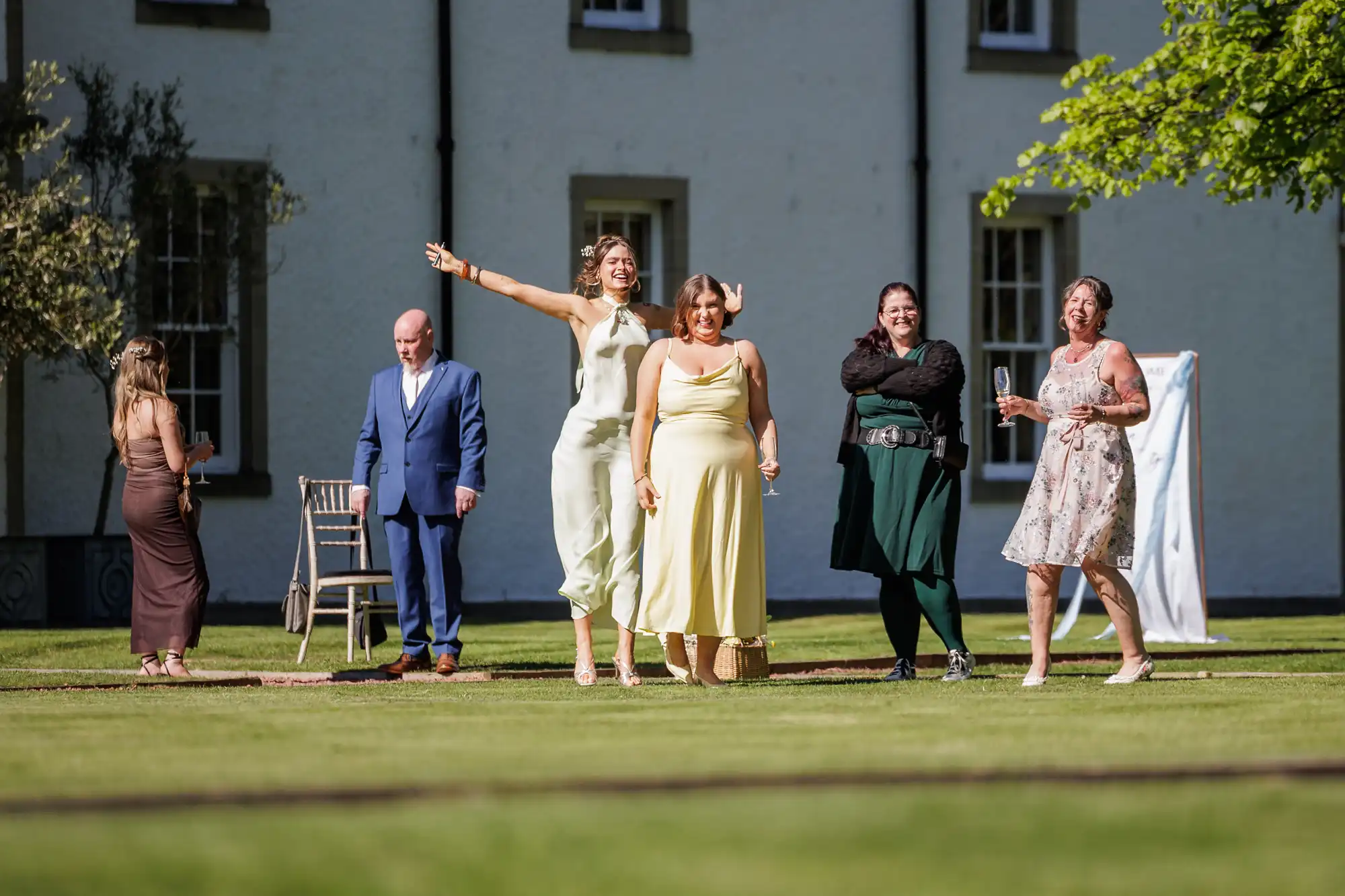 Five women and one man stand on a sunny lawn in front of a white building. Two women in pale dresses are laughing, one with arms raised. Others smile or watch. The man in a blue suit stands apart, looking down. Shadows and trees frame the scene.