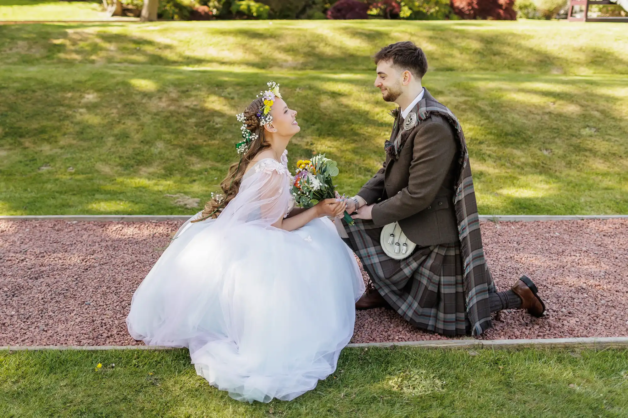 A bride in a white gown and flower crown kneels on a path, holding hands with a groom in a traditional Scottish kilt. Both are smiling at each other, outdoors on a green lawn with sunlight and trees in the background.