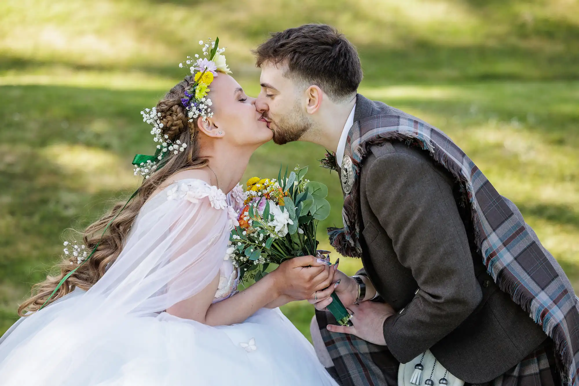 A bride in a white dress and flower crown and a groom in a tartan sash and brown suit gently kiss, holding a colourful bouquet, outdoors on a sunlit green lawn. Both appear joyful and dressed in wedding attire.