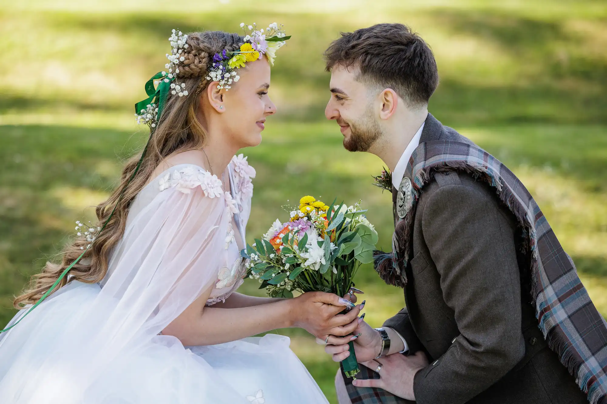 A bride in a floral crown and white dress holds a bouquet, smiling at a groom in a brown suit with a tartan sash. They kneel close, gazing at each other outdoors on green grass, sharing a joyful, intimate moment.