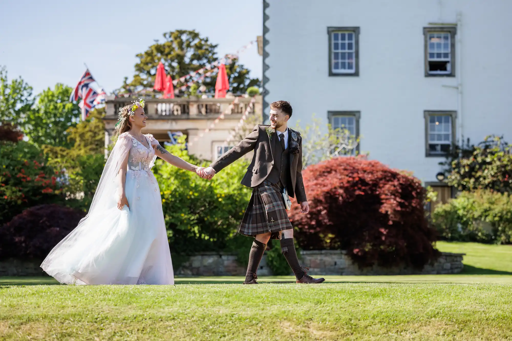 A bride in a white gown and flower crown holds hands with a groom in a dark jacket and tartan kilt as they walk on green grass outside a white building, with red umbrellas and UK flags visible in the background. Both are smiling joyfully.