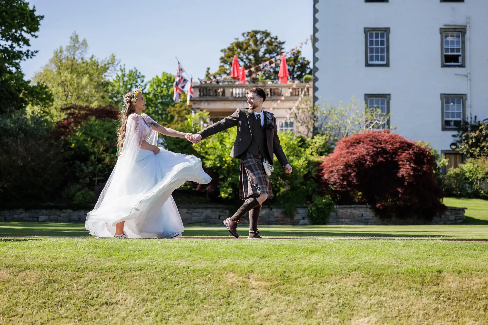 A bride in a flowing white dress and floral crown holds hands with a groom in a kilt as they joyfully walk on green grass, with an historic building, colourful trees, and British flags in the background on a sunny day.