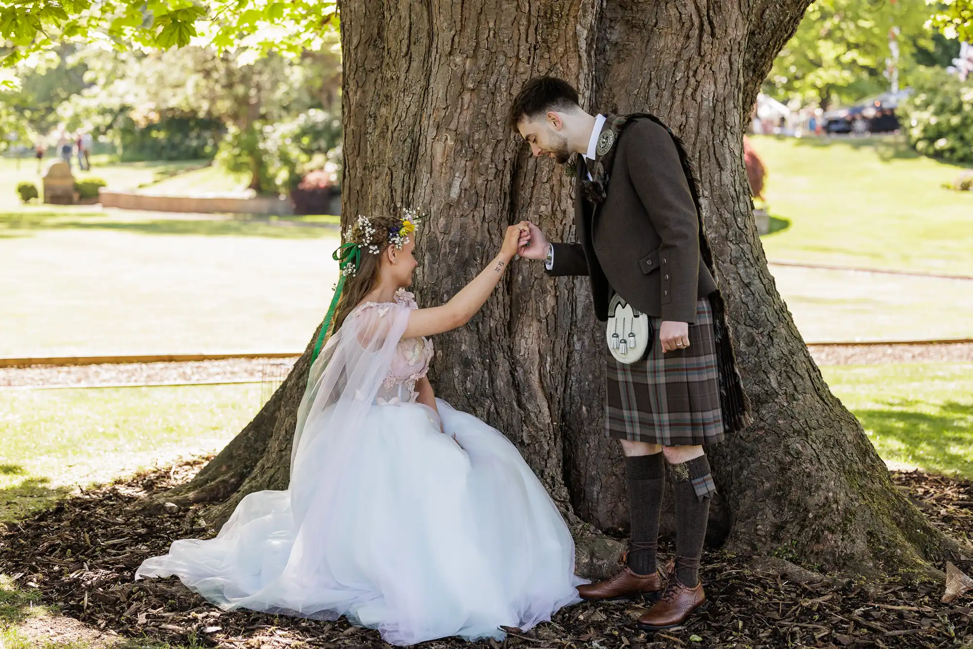 A bride in a white dress and flower crown sits under a large tree, holding hands with a groom in a brown jacket and tartan kilt, who stands leaning towards her. Sunlight filters through green leaves, and the scene is set in a grassy outdoor park.