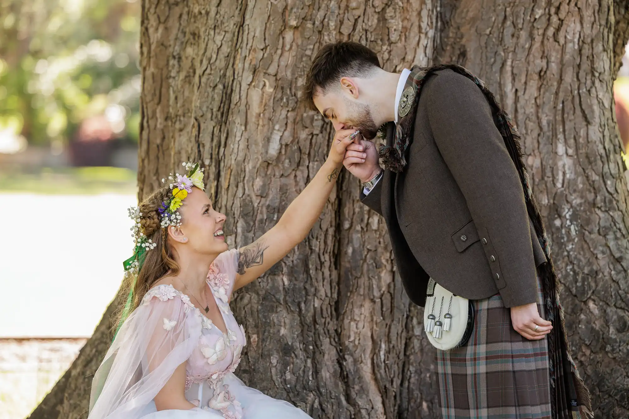 A woman in a floral crown and pink fairy dress kneels by a large tree, smiling as a man in a kilt and jacket bows and kisses her hand. Sunlight filters through the leaves, creating a whimsical, romantic outdoor scene.