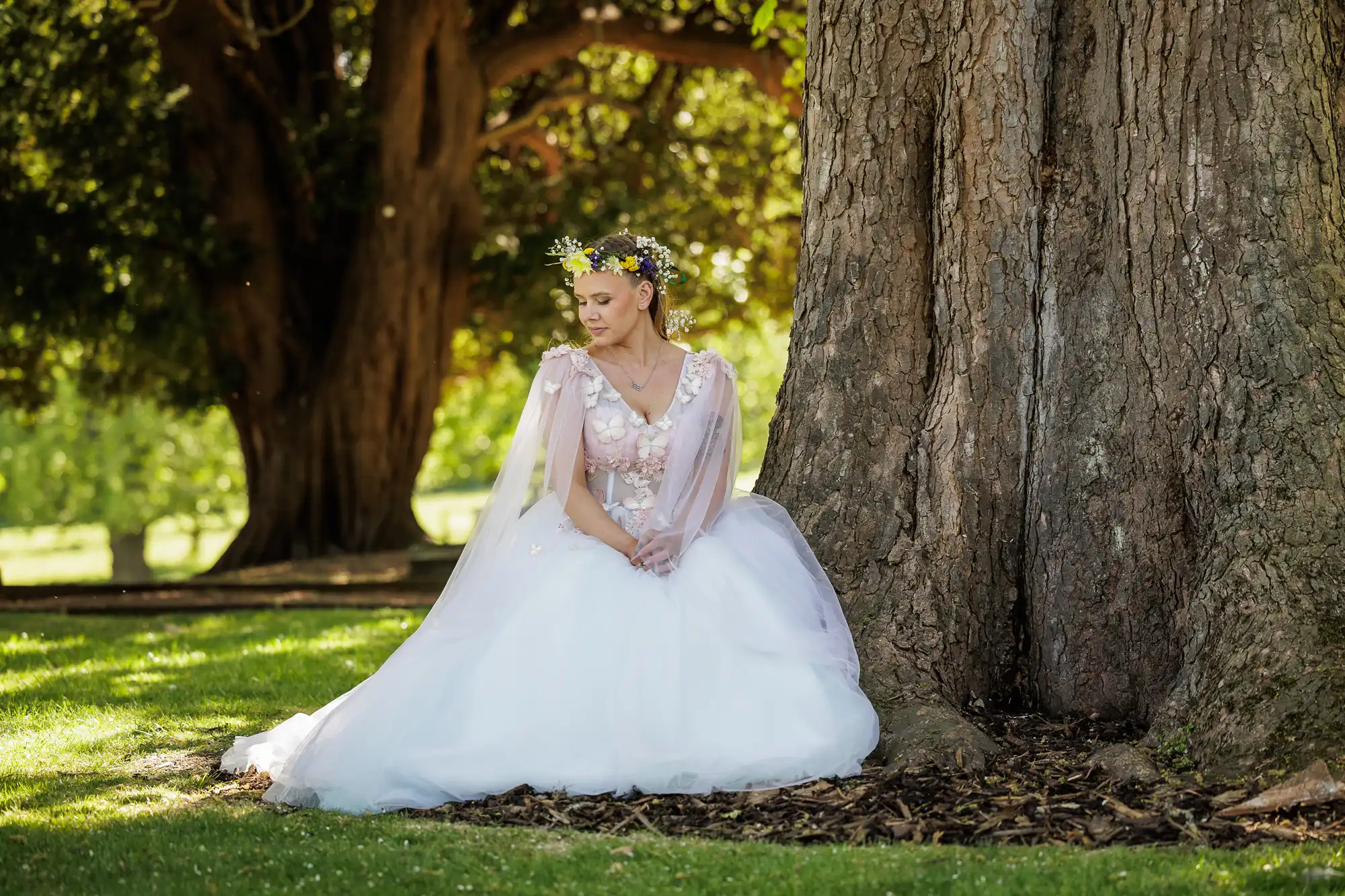 A woman in a white, flowing gown with sheer sleeves and floral details sits on the grass, leaning against a large tree trunk. She wears a flower crown and gazes down, surrounded by dappled sunlight and greenery in a peaceful outdoor setting.