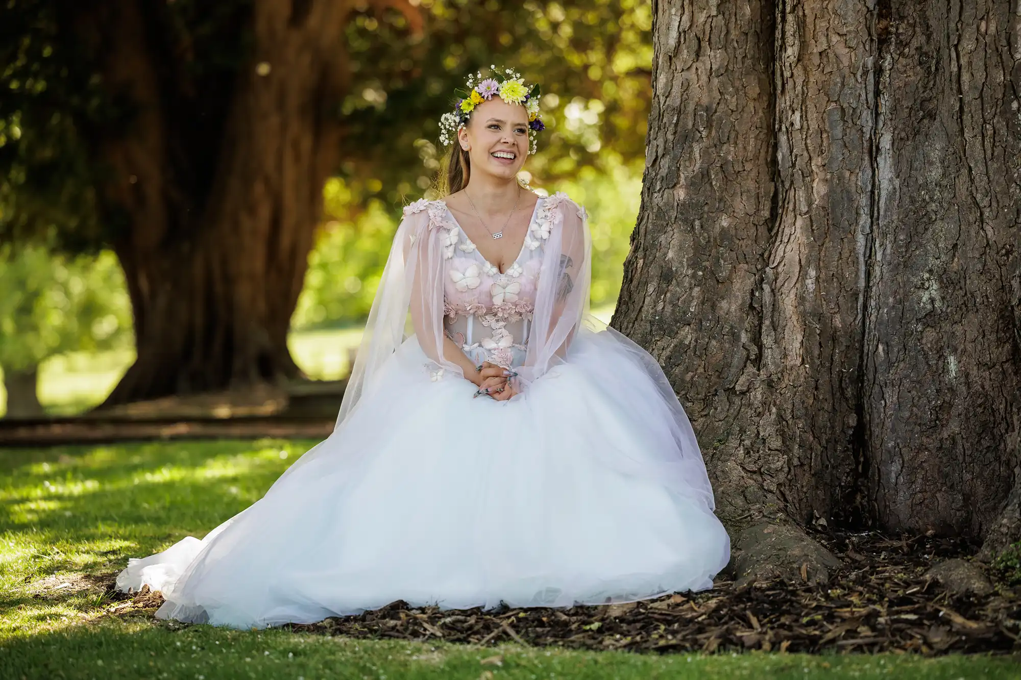 A smiling woman in a white, flowing gown and flower crown sits on the grass, leaning against a large tree in a sunlit park. Sunlight filters through the trees, creating a peaceful, fairy-tale atmosphere.