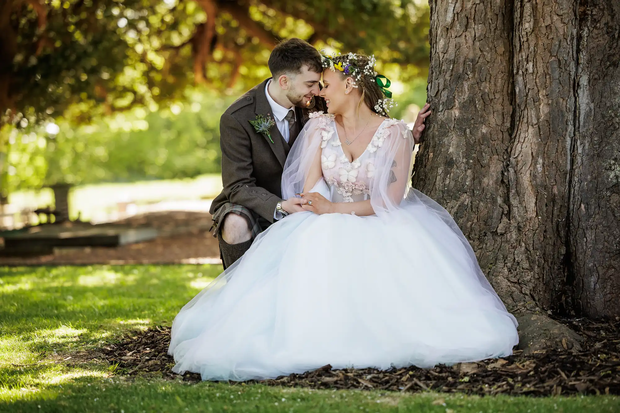 A bride in a white dress and flower crown sits with a groom in a brown suit under a large tree, touching foreheads and holding hands, surrounded by sunlight and greenery in an outdoor setting.