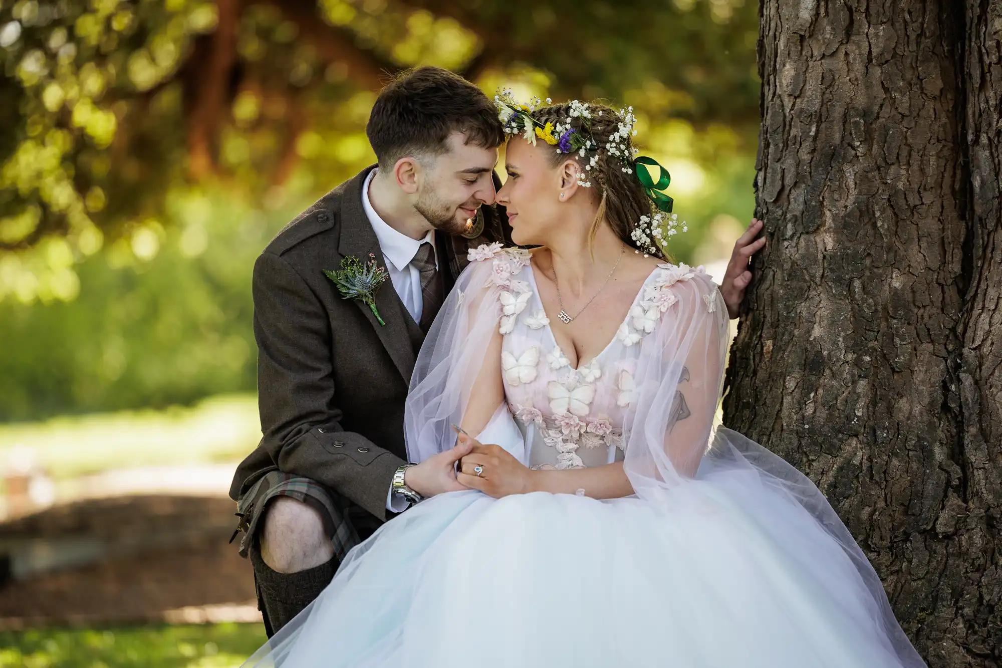 A bride in a white floral gown and flower crown sits by a tree, holding hands and gazing lovingly at a groom in a brown suit with a kilt. Sunlight filters through green leaves, creating a romantic and dreamy outdoor wedding scene.
